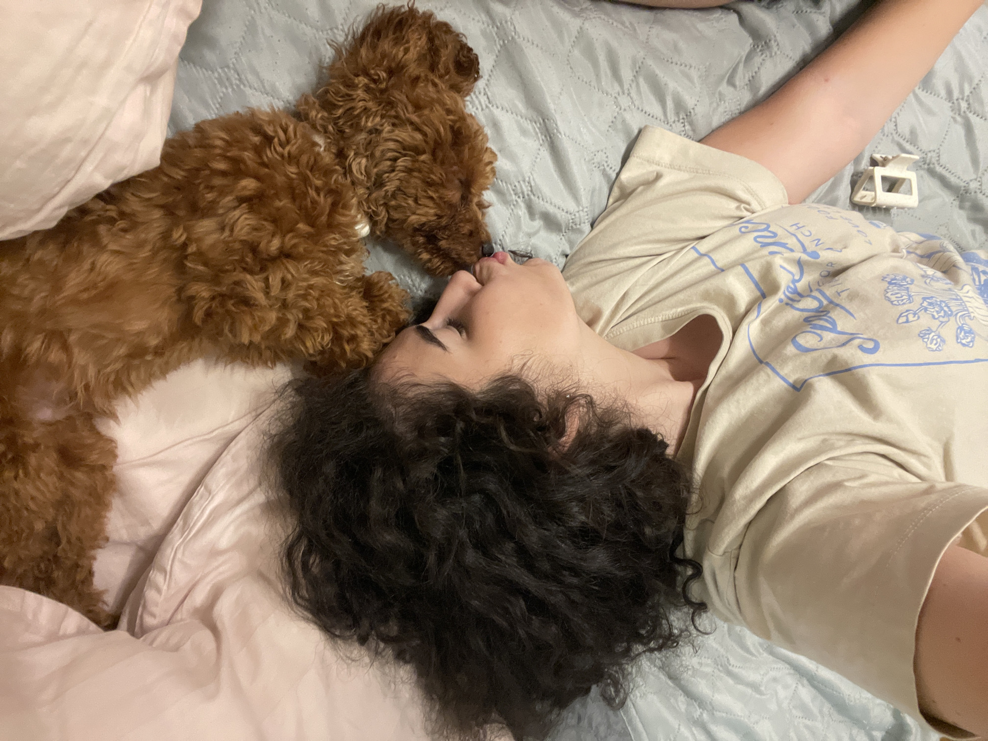 A person with dark curly hair and a light-colored t-shirt is lying on their side on a bed, facing a small brown dog. The person's eyes are closed and their lips are puckered, as if kissing the dog. The dog, a poodle mix, is also lying on its side, facing the person, with its head near the person's face. The bed is covered with a light blue quilted comforter. A pink pillow is visible on the left side of the image. The scene appears to be indoors, likely a bedroom. There is no information available about the location in Sofia, Bulgaria.