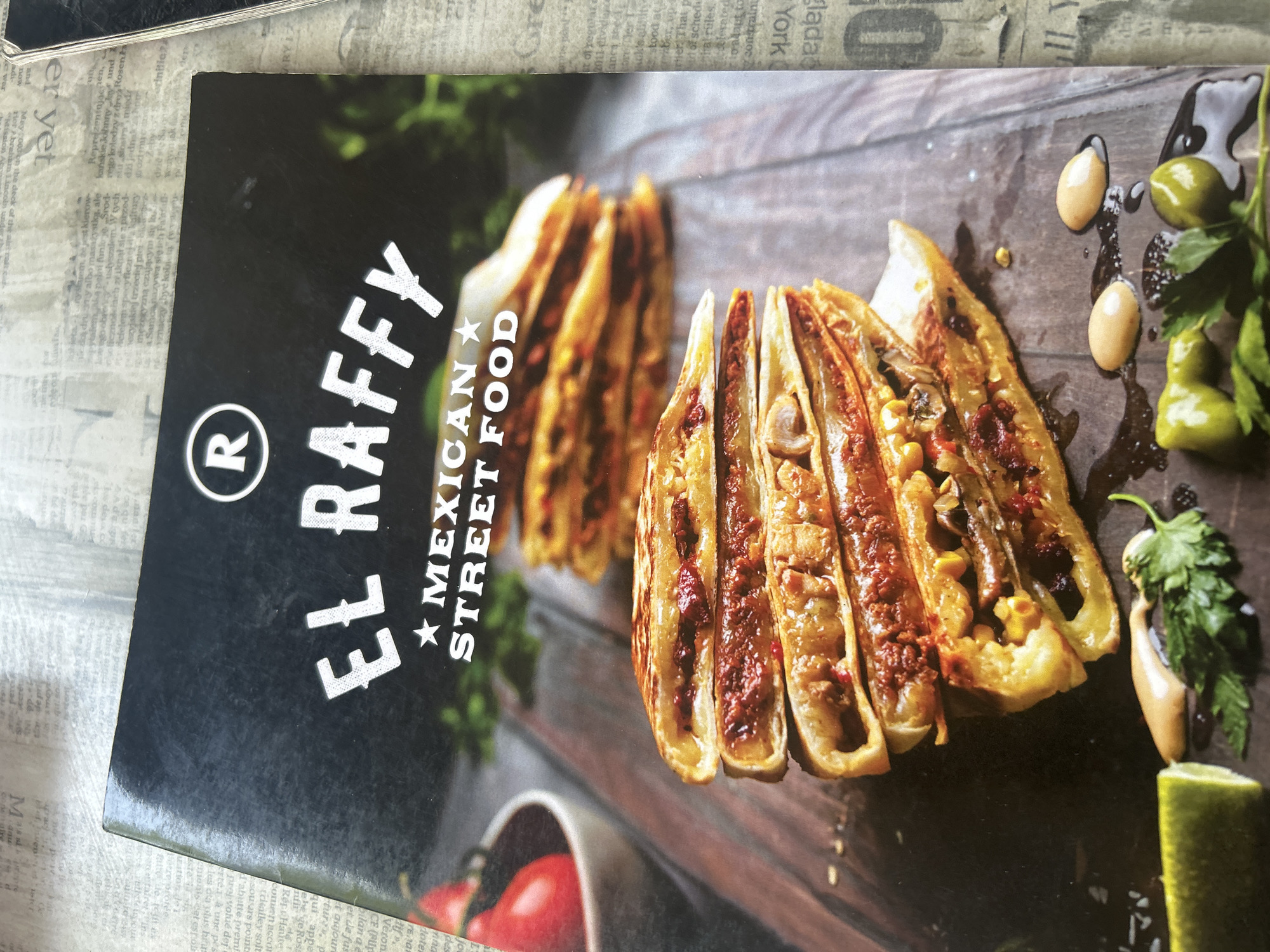 The image is a close-up shot of a menu or flyer for "EL RAFFY MEXICAN STREET FOOD." The background appears to be a rustic wooden surface. In the foreground, there is a stack of three quesadillas or similar folded tortilla dishes, cut in half to reveal the filling. The fillings appear to be a mixture of meat, vegetables like corn and mushrooms, and a red sauce. To the left of the quesadillas, there is a slice of lime and some fresh cilantro. To the right and slightly below the quesadillas, there are three dollops of a light-colored sauce and a few green olives. The flyer itself has the restaurant's name in large, stylized white font across the top, with a circular logo containing the letter "R." The overall presentation is an advertisement for Mexican street food. There is no information in the image that indicates a location in Plovdiv, Bulgaria.