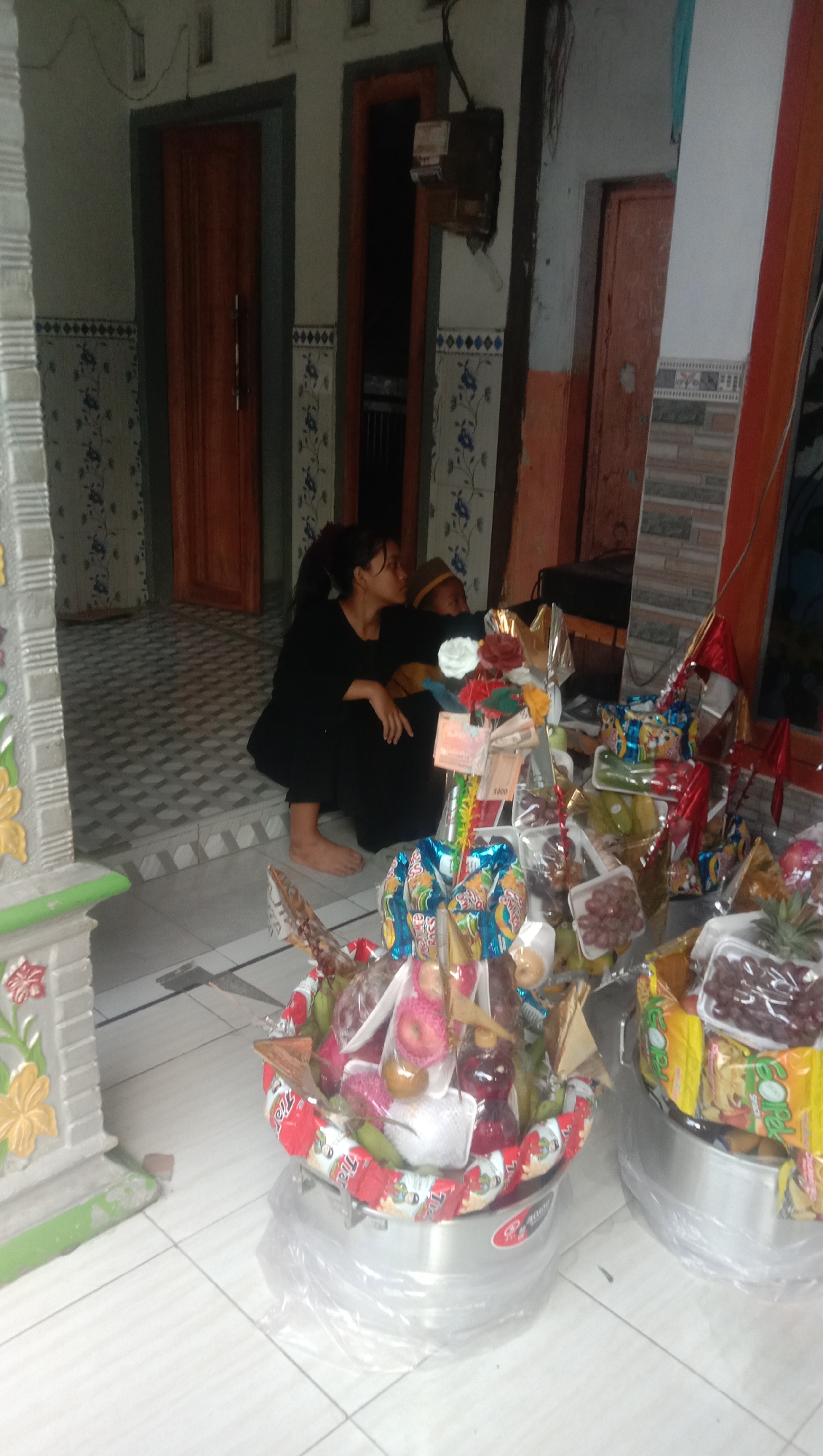The image depicts a scene on the floor of a building, likely a home, in Karang Anyar, Indonesia. In the middle ground, a woman in a black outfit is crouched down, facing to the right, with her left hand resting on her knee. Beside her, a child wearing a yellow cap is also partially visible. In the foreground, two large, round metal baskets are filled with various items. These baskets are decorated with colorful wrappers, snacks, fruits such as apples and grapes, and what appear to be small bottles of liquid and money. The baskets are covered in clear plastic. The background shows part of a building's interior with wooden doors, tiled walls with blue floral patterns, and an electrical box mounted on the wall. The floor is tiled with a checkerboard pattern.