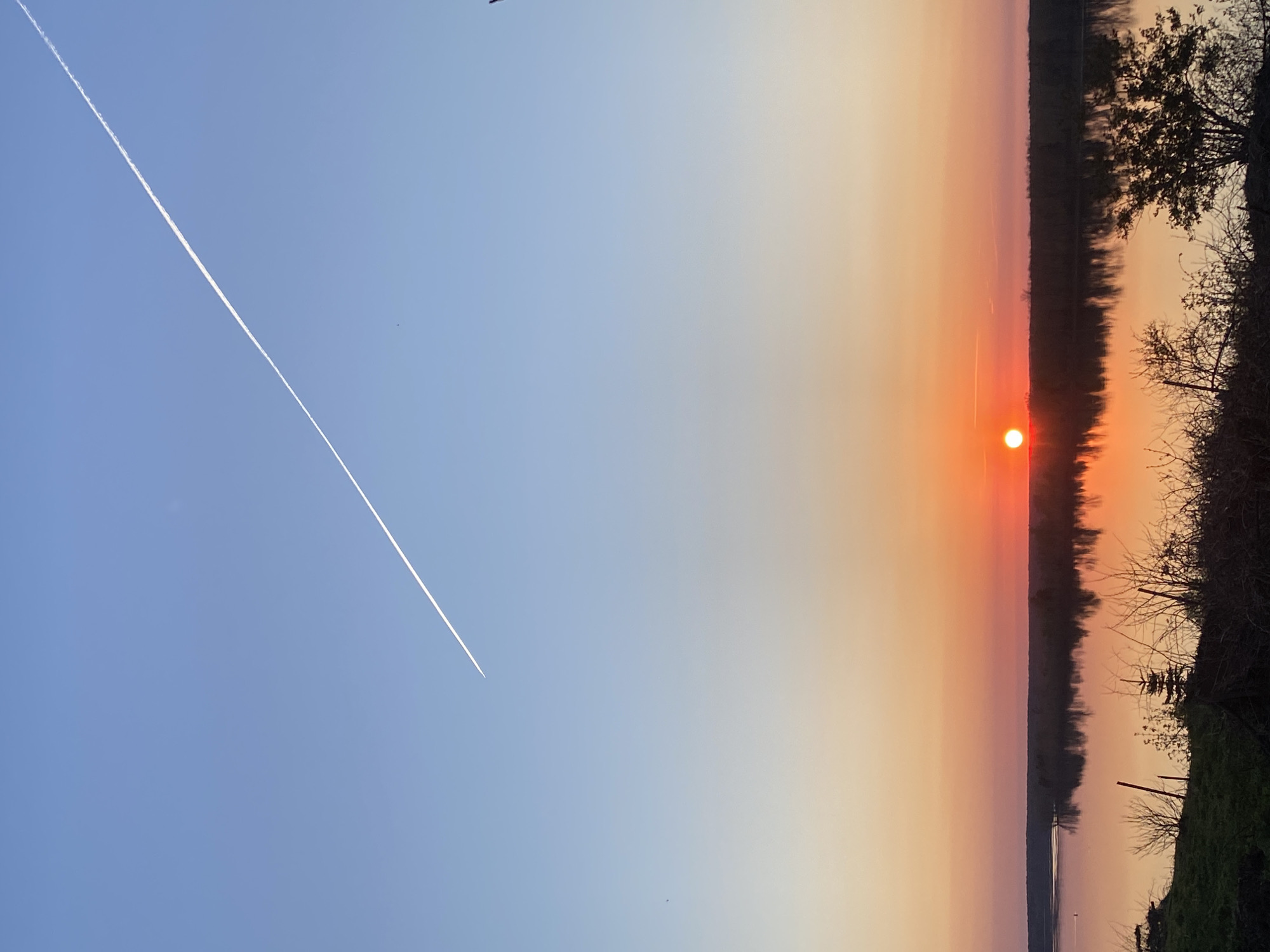The image captures a sunset over what appears to be a body of water or a river. The sky transitions from a deep blue on the left side to a gradient of orange and pink near the horizon. The sun is a bright orange sphere. Trees and vegetation are visible on the right side. A white contrail from an airplane cuts diagonally across the blue portion of the sky. The image orientation is rotated, with the sky positioned on the left and the horizon on the right. Ольховка, Russia, is not explicitly confirmed as the location.