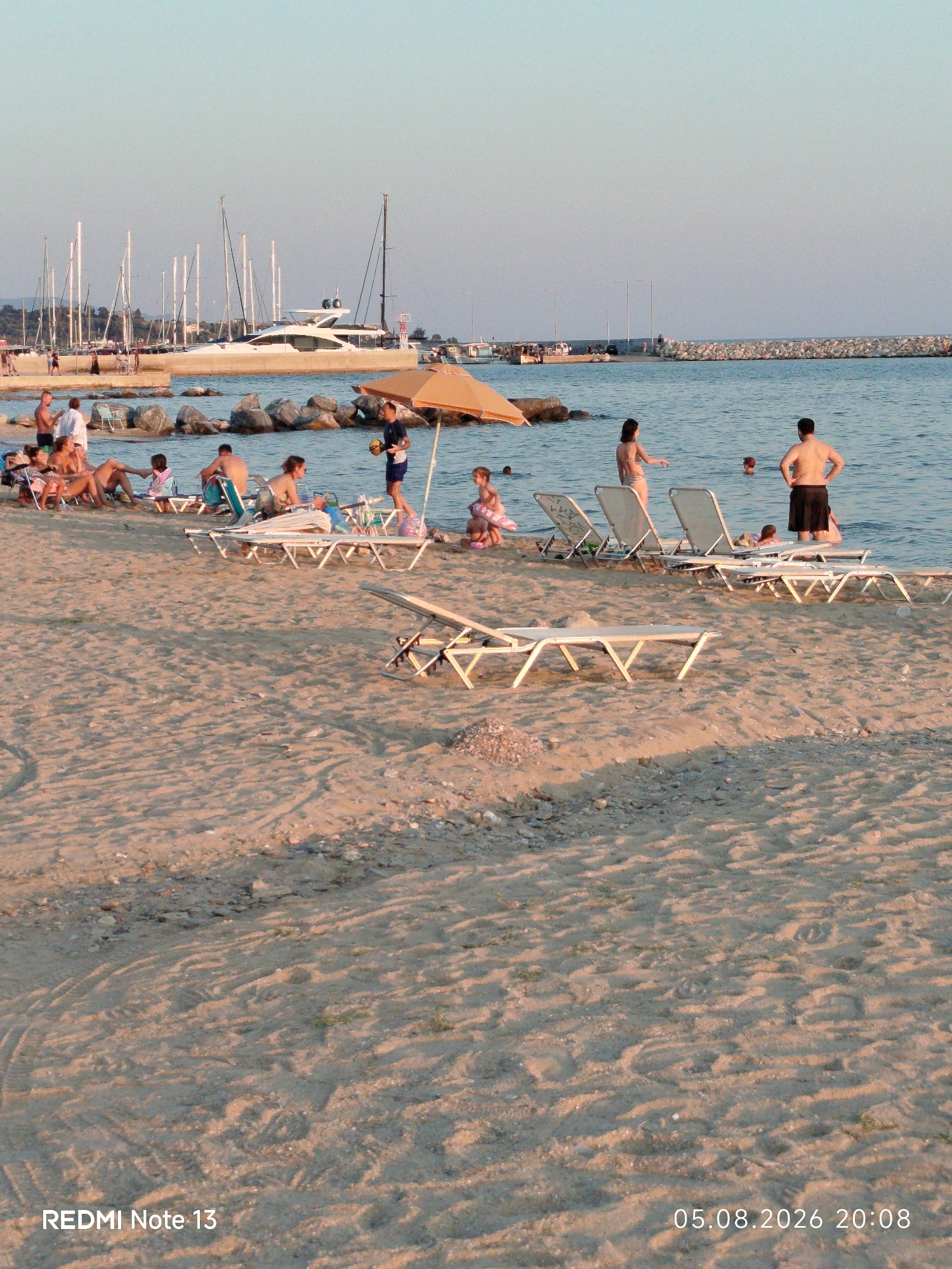 In Sithonia Municipal Unit, Greece, a sandy beach is populated by numerous individuals enjoying a sunny day by the sea. In the foreground, several empty lounge chairs are placed on the sand, suggesting a relaxing atmosphere. Further down the beach, groups of people are gathered, some sitting on chairs and others in the shallow water. A man in black shorts stands in the water, holding a round object, possibly a ball, while a woman in a bikini faces him. A child wearing a pink inflatable ring is visible near the water's edge, and other children are also present in the sea.
In the mid-ground, a large white yacht is docked at a harbor, with several sailboats moored behind it. A breakwater, constructed from large rocks, extends into the sea, separating the beach from the open water and the harbor. The sky is clear and pale blue, with the sun casting long shadows on the sand, indicating it is late afternoon or early evening. The overall scene depicts a typical summer day at a popular coastal resort.