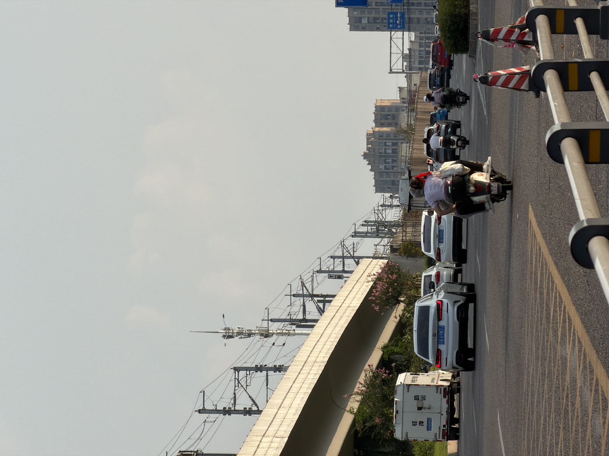 In Jiangjin, China, on a clear day, a street scene unfolds with multiple vehicles on a paved road. Overhead, an elevated structure with train tracks and numerous utility poles stretches across the frame, indicating proximity to a transportation hub. In the foreground, traffic cones with red and white stripes mark a barrier. Cars, including a white Audi, are seen in traffic. Several people are riding on motorcycles and scooters, with one scooter carrying two passengers. In the background, apartment buildings and a communication tower are visible. The overall environment suggests an urban setting with active road traffic and infrastructure.