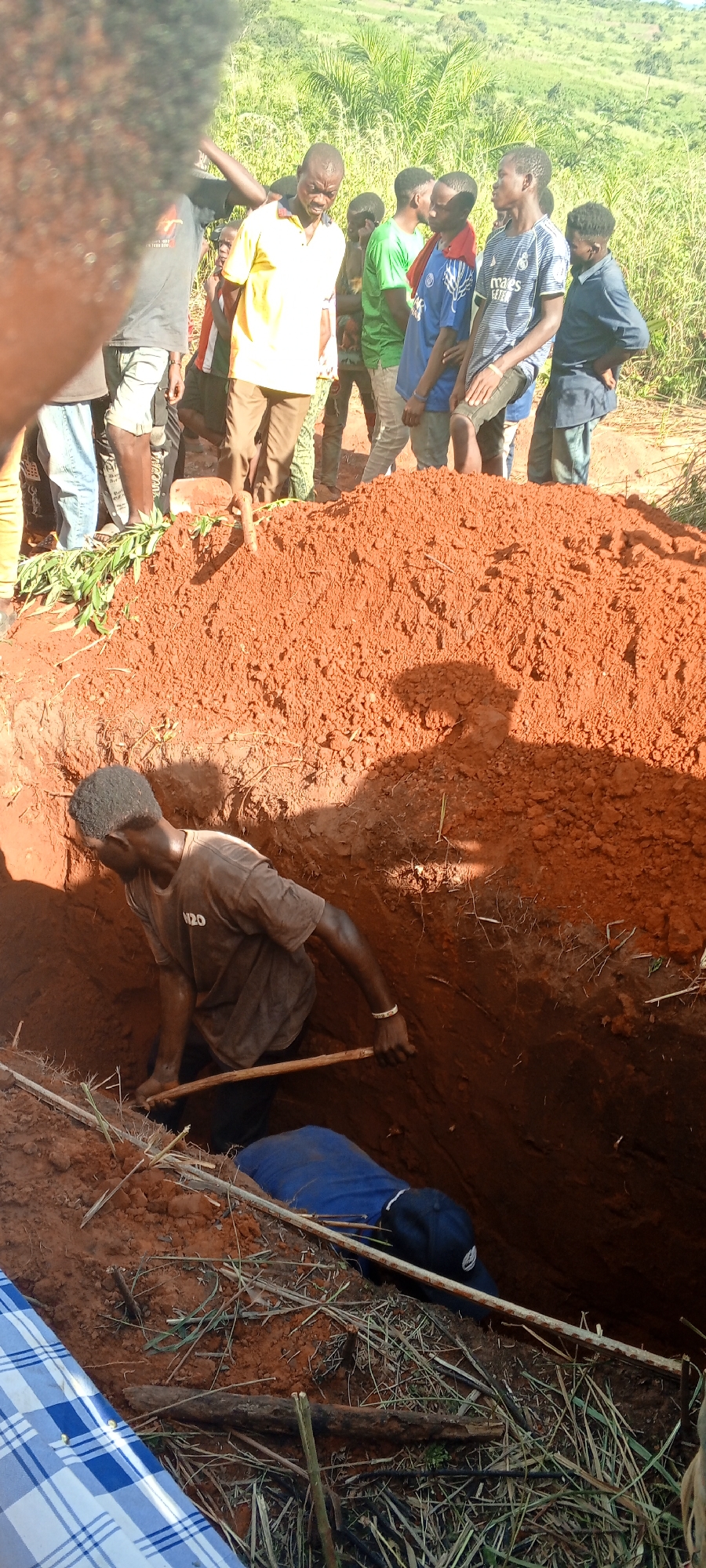 In Ngalula, Democratic Republic of the Congo, a man is digging a grave in a reddish-brown dirt pit. He wears a dark gray t-shirt and black pants, and holds a wooden stick. Inside the grave, a body wrapped in blue fabric is partially visible. A group of people stand around the grave, observing. The background shows green vegetation and a hillside.