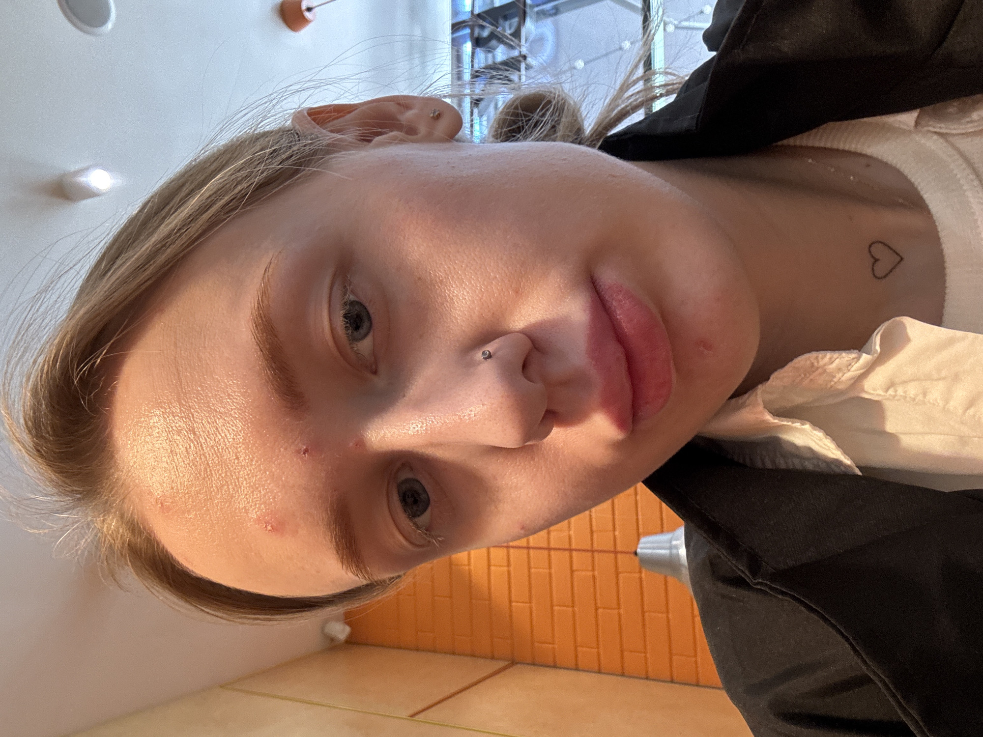 A close-up, slightly tilted selfie shows a young, fair-skinned person with blonde hair pulled back, revealing a pierced nose and ear. The individual has a heart-shaped tattoo on their upper chest/neck area. A black jacket is visible. The background includes what appears to be an orange brick wall or tiling and a glimpse of a room with vertical bars or a glass structure. The location in Rostov-on-Don, Russia, is not determinable from the image.