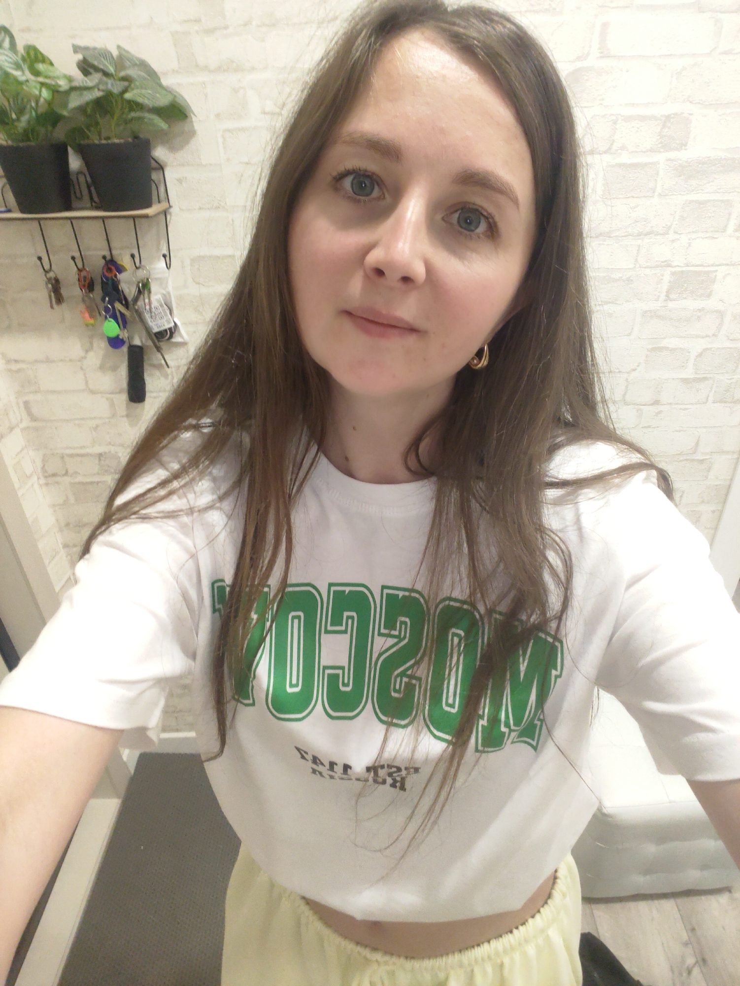 A young woman with light brown hair and blue eyes is centered in a selfie, wearing a white T-shirt with "MOSCOW" printed in green lettering. She also has a small gold hoop earring on her left ear. The background features a white brick-patterned wall, on which hangs a black wire rack holding two small potted green plants and a collection of keys and other small items. A gray-toned flooring is visible at the bottom of the frame, and a white object is seen to the right. The bottom of the frame also contains her lower waist.