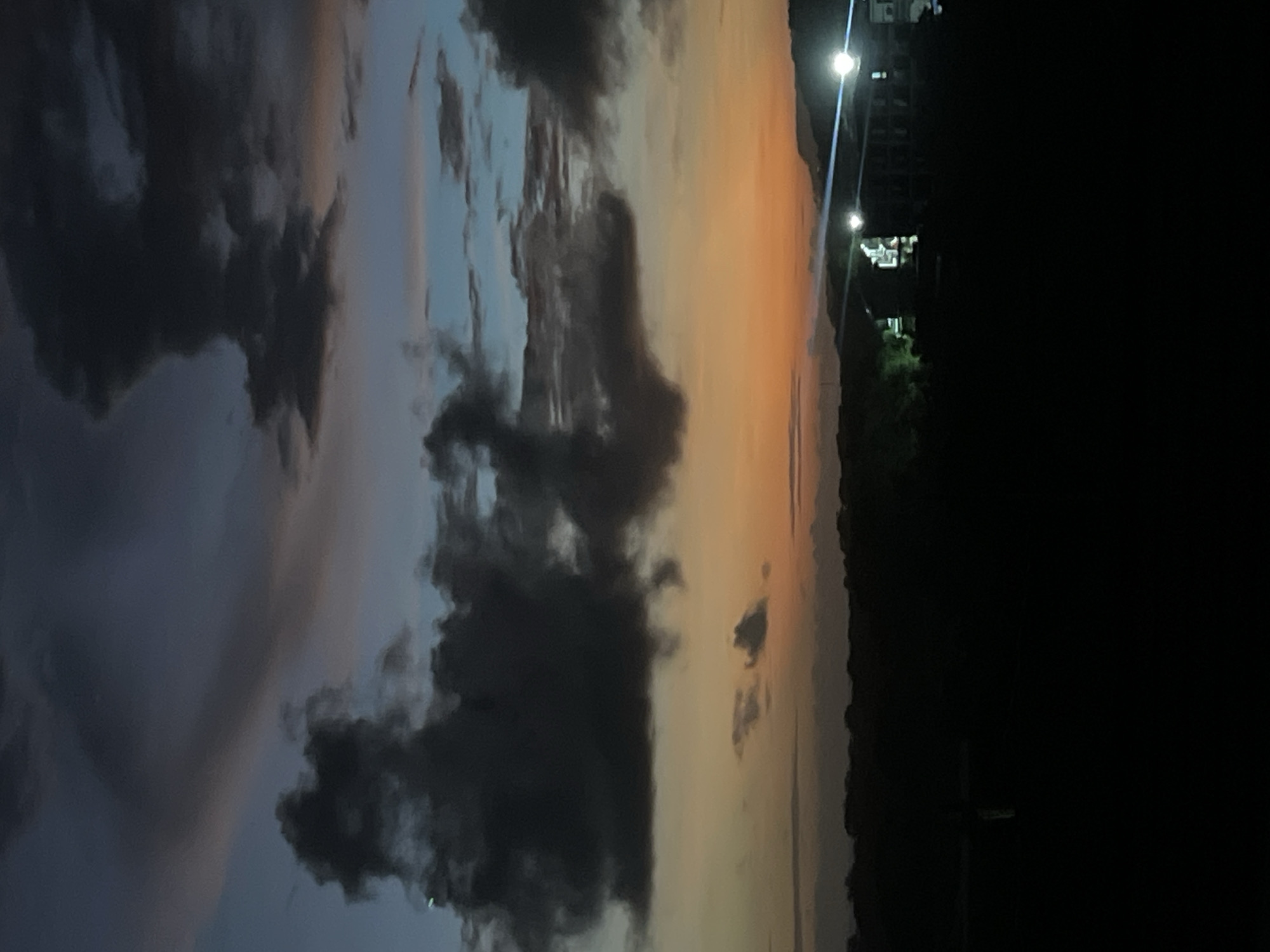 The image depicts a night scene with a view of the sky and a dark foreground. The sky displays a gradient of colors, transitioning from orange near the horizon to blue higher up, with dark clouds scattered throughout. On the right side of the image, there is a dark area, possibly representing a landscape or buildings, with faint lights visible. There are no people visible in the scene. The environment appears to be a city, given the presence of lights and buildings in the distance, but the specific location is not identifiable from the image.