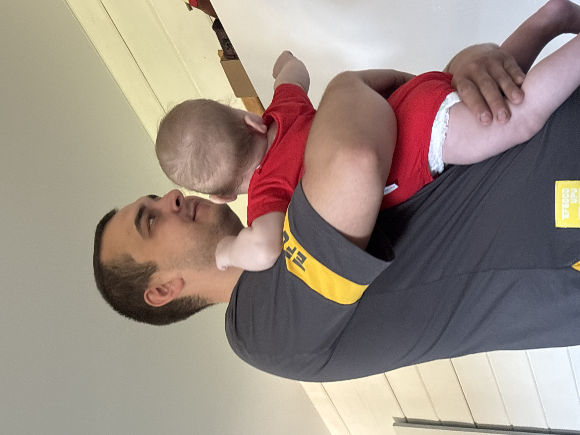 A man is holding a baby. The baby is wearing a red outfit and a diaper. The man is wearing a gray shirt with yellow trim on the sleeve. The location is unknown, and there is no information to determine the city of Раздолье, Russia.