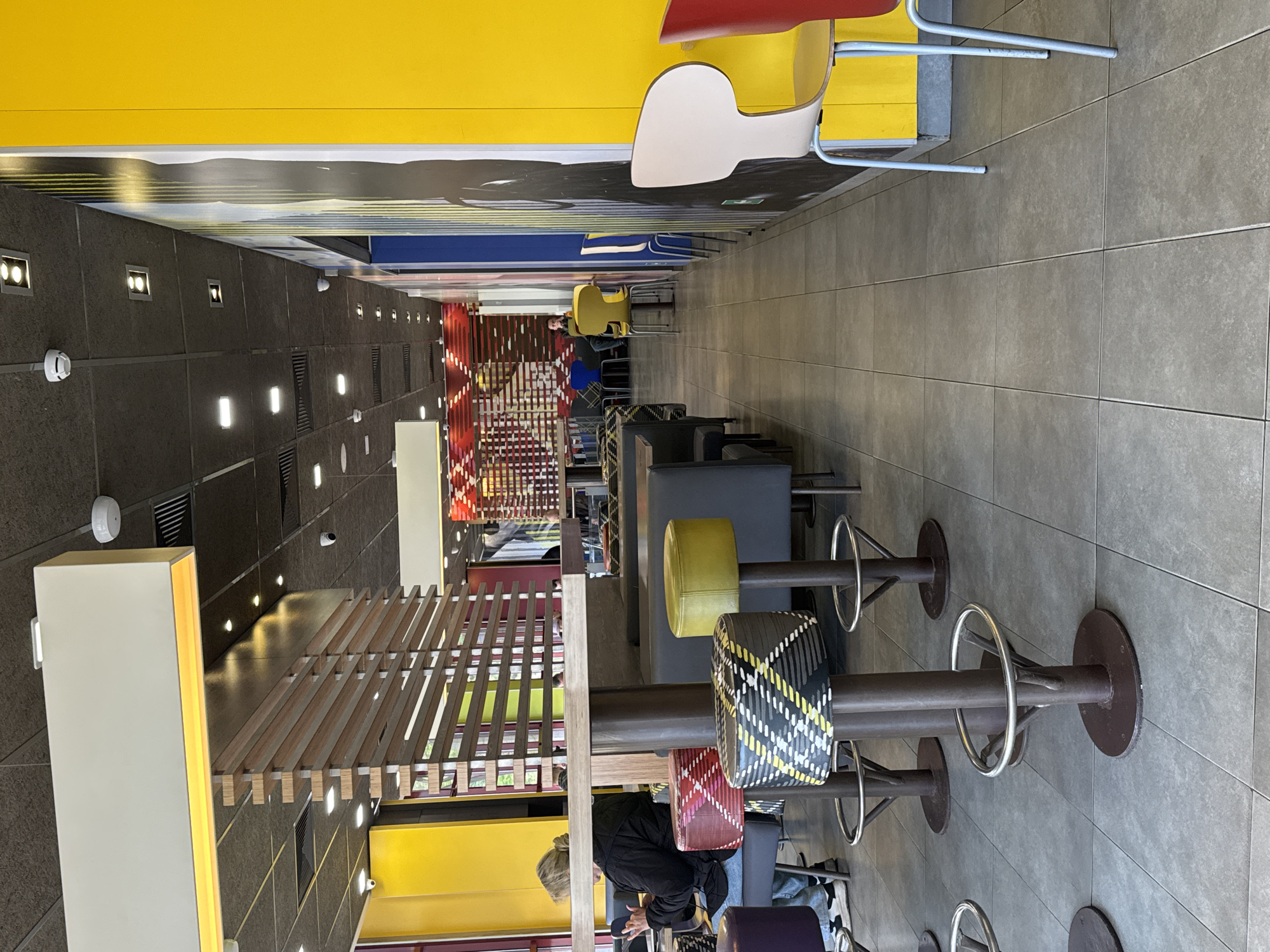 The image depicts the interior of a McDonald's restaurant. The scene is shot from a high vantage point, looking down a long, narrow space filled with tables and high-top chairs of various colors. Patrons are seated and eating at the tables. The interior design features yellow and black elements along with light wood slats. The floor is tiled in a neutral grey color. The lighting is provided by recessed lights along the walls and ceiling. The image suggests that the photograph was taken in Saint Petersburg, Russia.