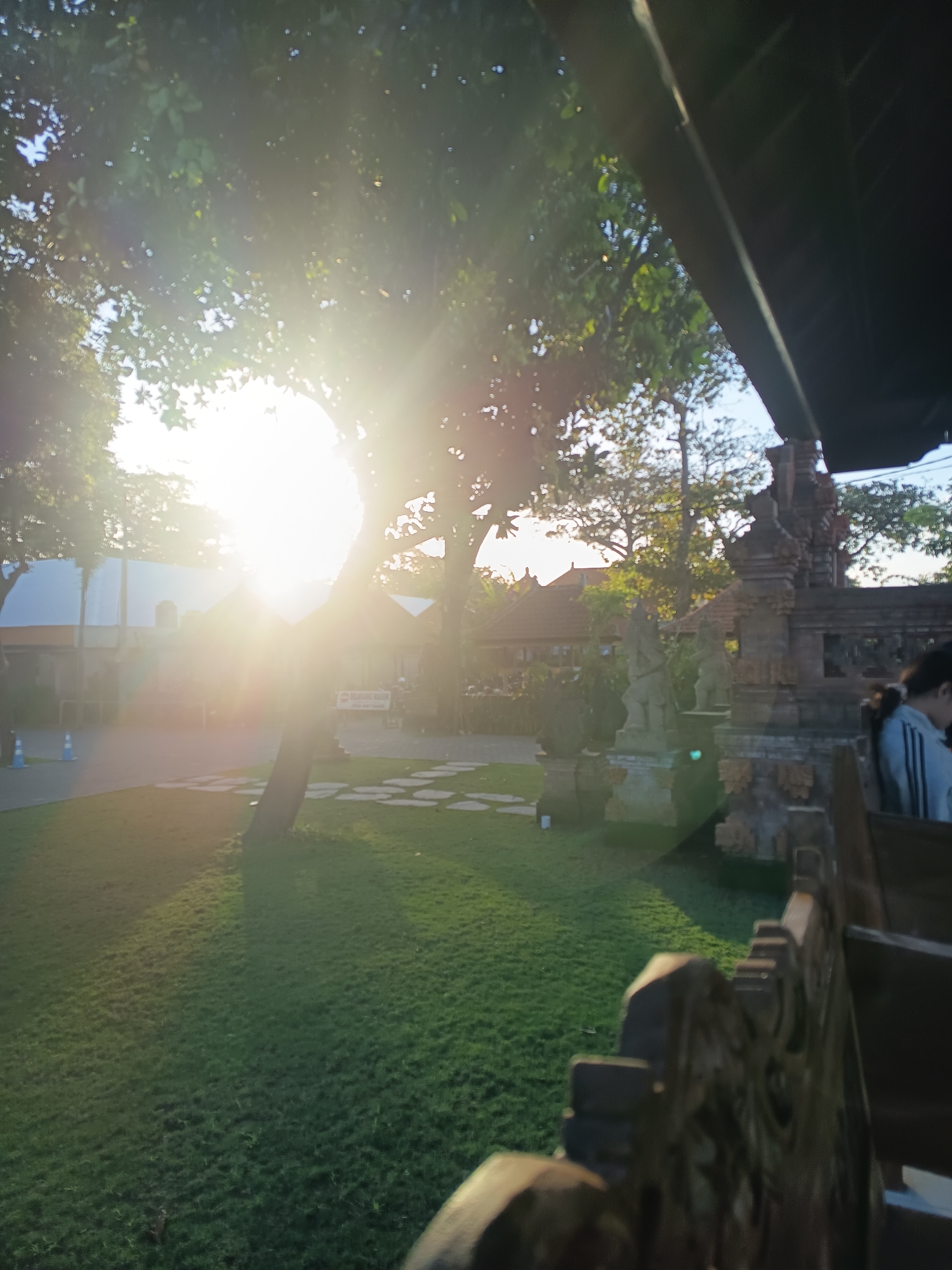 The image depicts a sun-drenched outdoor scene, likely a park or courtyard in Denpasar, Indonesia. The sun, a prominent bright white orb, is shining intensely through the branches of a large tree, creating strong lens flare and a radiant glow across the upper left portion of the frame. Green grass covers the ground in the foreground, with irregular stone pathways visible across it.
In the background, traditional Balinese architecture is evident. A carved stone gate or structure stands to the right, adorned with intricate details and statues. Behind this, buildings with terracotta tiled roofs and stone facades can be seen, suggesting a cultural or historical site. A person, partially visible on the far right, is seated, wearing a light-colored top with stripes. Another person is partially visible in the distance, near a white tent-like structure.
The overall atmosphere is bright and warm, indicative of a sunny day, possibly late afternoon given the angle of the light and the long shadows cast on the grass. The scene appears tranquil, with a blend of natural elements and cultural architecture.