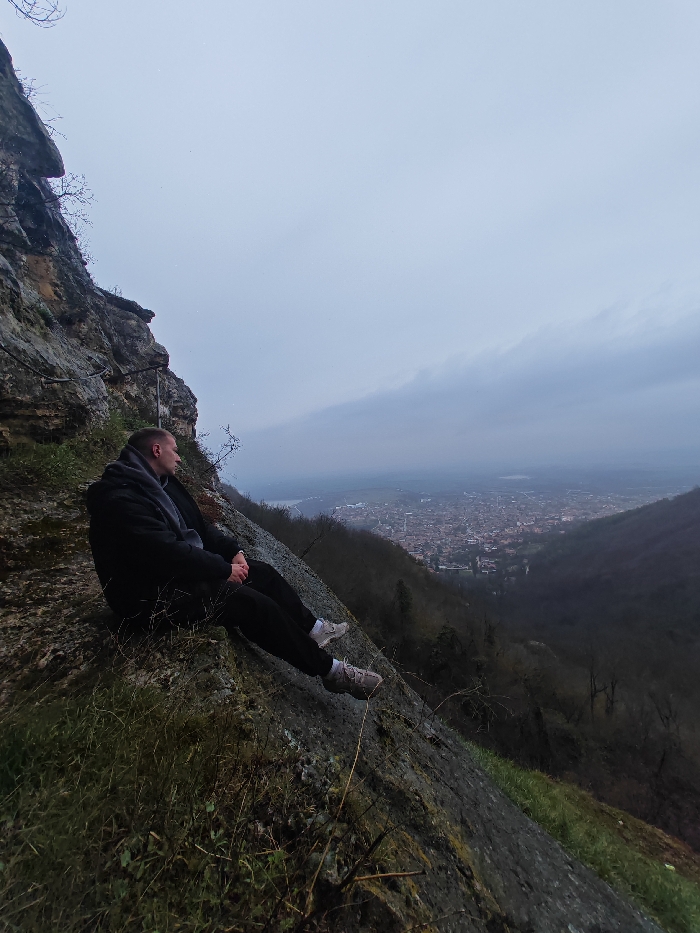 A lone male subject is observed seated in profile on a steep, rocky incline, part of the Shumen Plateau, overlooking the city of Shumen, Bulgaria. The individual wears a black jacket, a dark grey hooded garment or scarf, black trousers, and light grey athletic shoes with white soles. His hands are clasped in front, and he is gazing towards the sprawling cityscape in the valley below. The immediate foreground and midground consist of a weathered, textured rock face with sparse dry grass and leafless shrubbery. Metal cables or handrails are visible anchored to the rock behind the subject, indicating a maintained path. In the background, the urban area of Shumen is spread across a valley, flanked by hills covered in deciduous trees that appear bare. A body of water is discernible beyond the city. The sky is uniformly overcast and grey, suggesting a dull weather condition.
