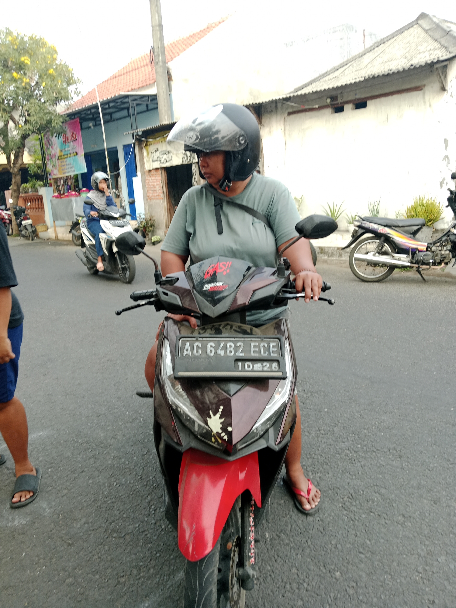 A person wearing a helmet and a green t-shirt is riding a motorcycle with a red front fender. The motorcycle has a license plate that reads "AG 6482 ECE" and "10/26". The person's right hand is on the handlebar. In the background, another person is riding a white motorcycle, and buildings and trees are visible. The scene appears to be a street in Kediri City, Indonesia.