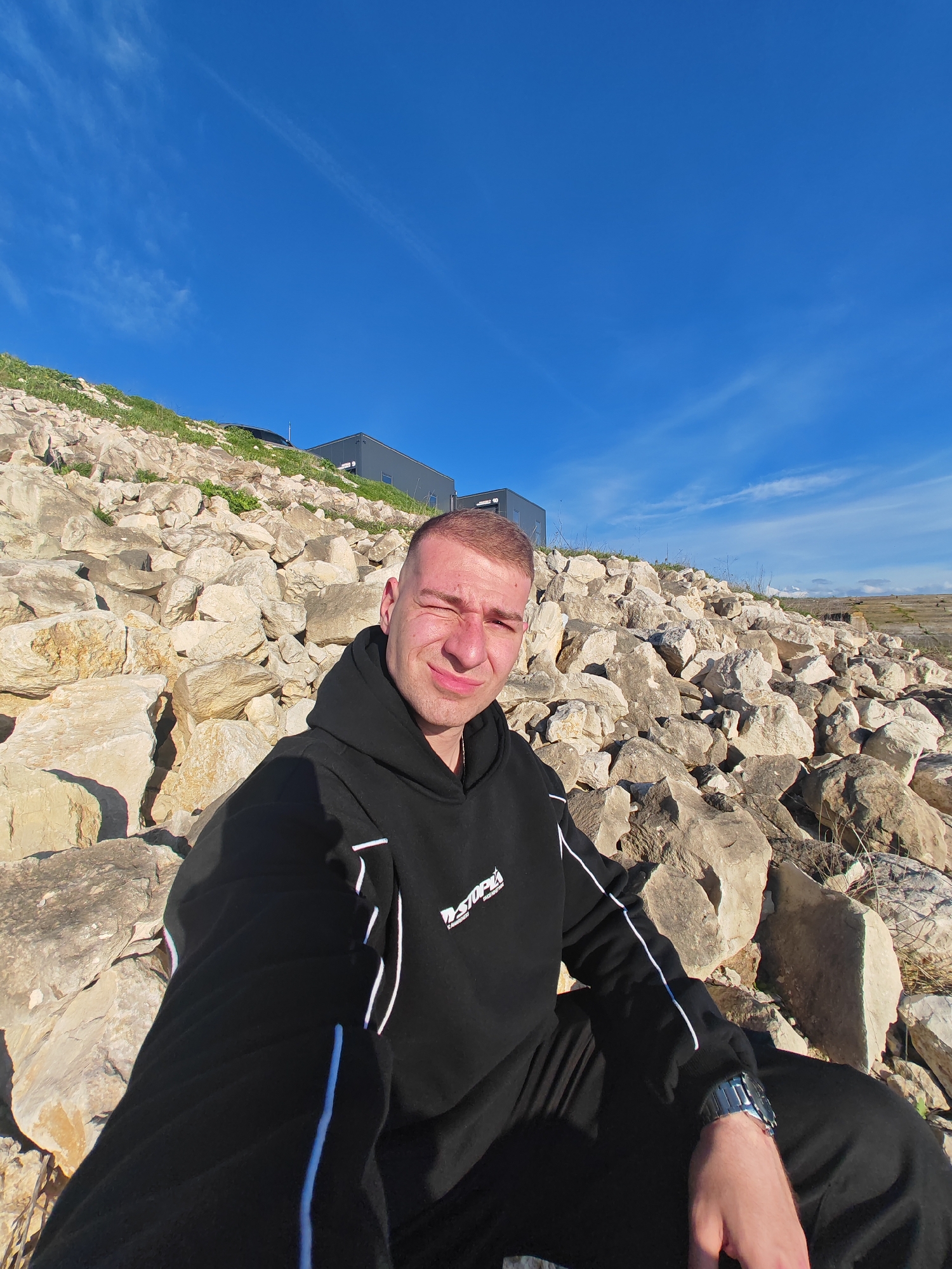 A man wearing a black tracksuit is sitting amongst large rocks in Shumen, Bulgaria. The building visible in the background is located at coordinates 43.27883, 26.92094. The sky is blue and partly cloudy.