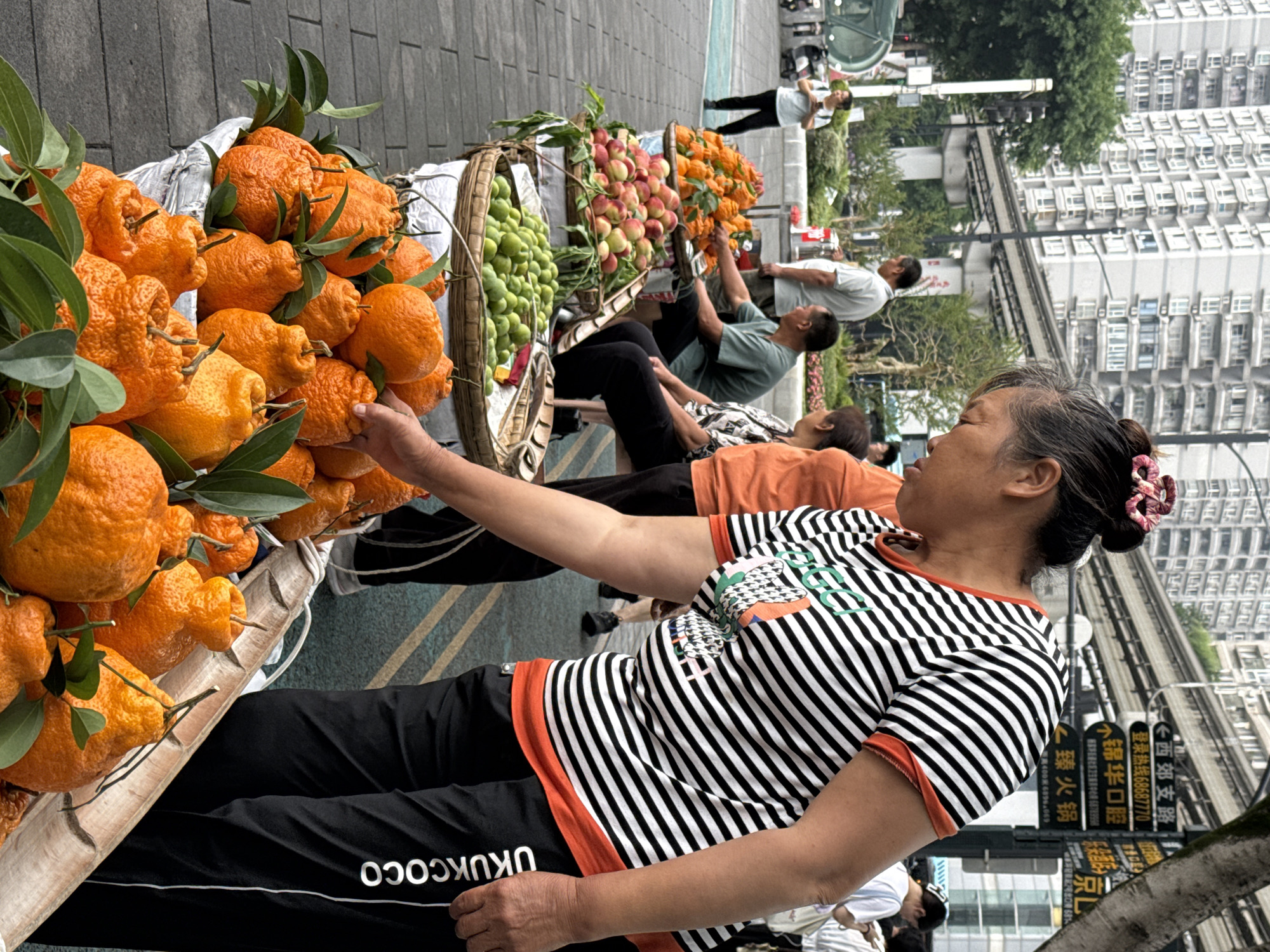 The image captures a scene in Jiulongpo District, China, portraying a street vendor selling fruit. The vendor, a woman with her hair styled in a bun adorned with a floral accessory, wears a striped shirt with orange accents and black pants featuring "UKUKCOCO" text along the leg. She is positioned next to a cart laden with bright orange mandarin oranges with green leaves still attached. A basket with green limes and another with red apples sit beside the oranges. The street has a few pedestrians, with a man in a white shirt observing the scene and another with an orange shirt sitting next to her.
In the background, modern architecture is visible, with buildings towering behind the vendor. Elevated light rail tracks run along the street, and street signs with Chinese characters are present. The environment appears to be a busy, urban setting with commerce taking place on the sidewalk.