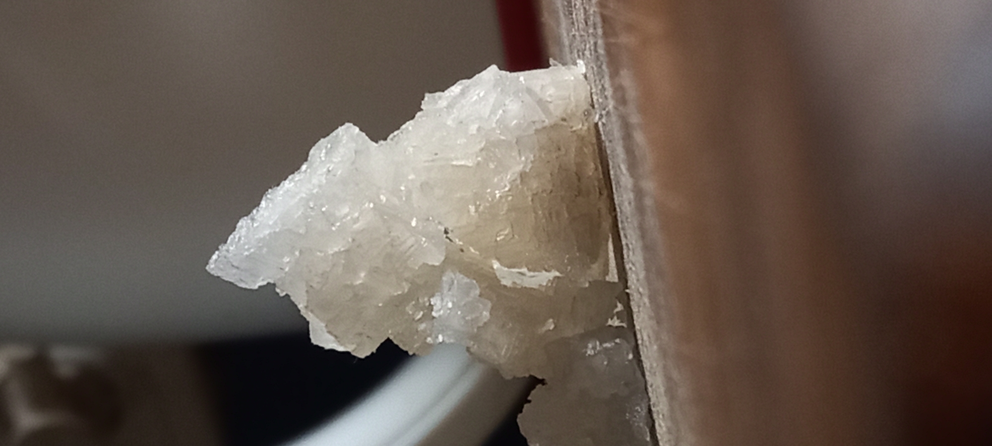 The image displays a close-up view of a white, translucent crystalline formation attached to the edge of a flat, brown, wood-like surface. The crystals exhibit an irregular, somewhat flaky, and granular texture, with individual crystalline structures visible. They appear to be growing outwards from the vertical brown surface, which could be a wooden beam, panel, or part of a piece of furniture.
In the background, a blurred greyish wall or surface is visible, and a small, blurred reddish area can be seen near the top left of the background. Below the crystalline formation, a blurred white object, possibly a cable or another structural element, is partially visible.
The scene suggests an indoor or sheltered environment. The appearance of the crystals could indicate either ice formation (such as frost or rime ice) due to cold temperatures, or mineral deposits (like efflorescence from salts or other soluble compounds) resulting from moisture evaporation. There are no human subjects or activities depicted. No text is visible in the image. The location context for this image is Kryvyi Rih, Ukraine.