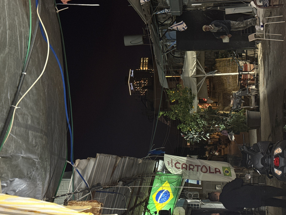 The image depicts a nighttime scene in an urban alleyway within Tel-Aviv, Israel. The background features the illuminated Kempinski Hotel Tel Aviv building, confirming the city location.
Overhead, a grey tarp awning spans the width of the frame, supported by wires and ropes, with multiple colored electrical cables (white, black, green, blue) running beneath it. On the left, a structure with corrugated metal panels and exposed wiring displays an illuminated Brazilian flag hanging vertically, along with a white banner bearing the Hebrew word "כשר" (Kosher) and "CARTOLA" in red text, accompanied by a small Brazilian flag symbol. A male subject, seen from the back in dark attire, stands near a dark scooter or motorcycle in the lower-left foreground.
The mid-ground right features potted plants, including a citrus tree with yellow fruits, and an open white patio umbrella surrounded by tables and chairs. Adjacent to this area is a dark vertical structure from which several small Israeli flags are hanging. A second male subject is visible on the right, wearing a light-colored beanie, a dark jacket, and light-colored, paint-splattered pants. He holds a white container or bucket in his left hand while appearing to interact with a dark vertical surface, possibly engaged in cleaning or painting. Two empty light-colored chairs are positioned in the immediate right foreground. The ground surface appears to be asphalt or concrete, with visible wet areas. The alley recedes into the darkness, with other dimly lit buildings in the distant background.