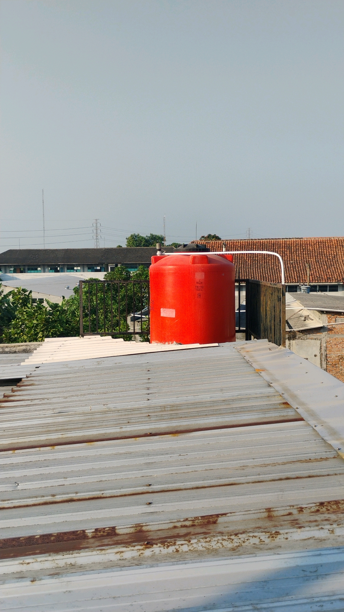 In Gumpang, Indonesia, a bright red water tank sits atop a weathered corrugated metal roof. The tank, cylindrical in shape, is positioned in the mid-right of the frame. Behind it, a brown tiled roof of a building is visible, along with a section of brick wall and a dark wooden fence. Green foliage from trees peeks out from behind a black metal railing situated to the left of the water tank. In the background, a long, dark-roofed building with visible windows stretches across the scene, partially obscured by trees. Power lines and transmission towers are discernible against a pale, hazy sky. The foreground is dominated by the textured surface of the rusted corrugated metal roof, its unevenness and signs of wear prominent. The scene suggests an urban or semi-urban environment, with residential or commercial structures. No people are present in the image.