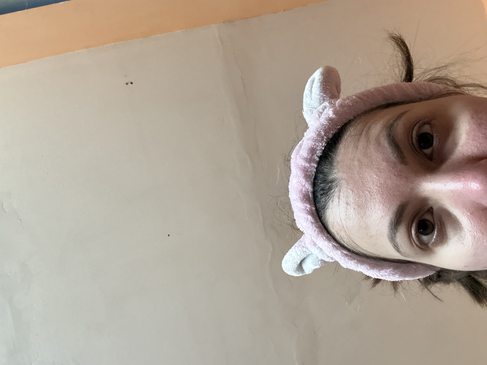 A close-up, tilted, upward-angle shot shows a person with fair skin, brown eyes, and dark hair, wearing a pink and white fuzzy headband with ear-like protrusions. The person's face is partially visible, showing the forehead, eyebrows, eyes, nose, and part of the cheek. The background is a light beige or tan wall, with a darker orange or peach-colored section visible in the upper left corner. There are also a couple of small dark spots on the wall. The location is unknown.
