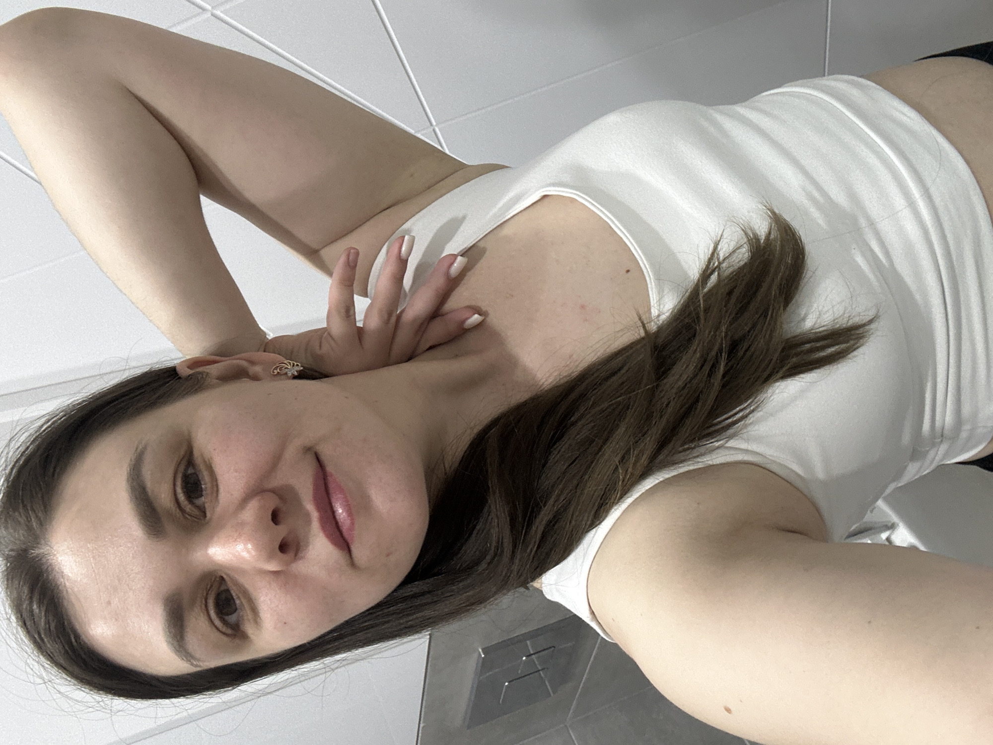 A woman is posing for a selfie in what appears to be a bathroom. She has light skin, brown hair, and is wearing a white tank top. Her left arm is raised behind her head, and her right hand is near her neck. The background consists of white tiled walls and a gray toilet flush plate. There is no verifiable evidence to confirm the location as the city of Chaikovsky, Russia.
