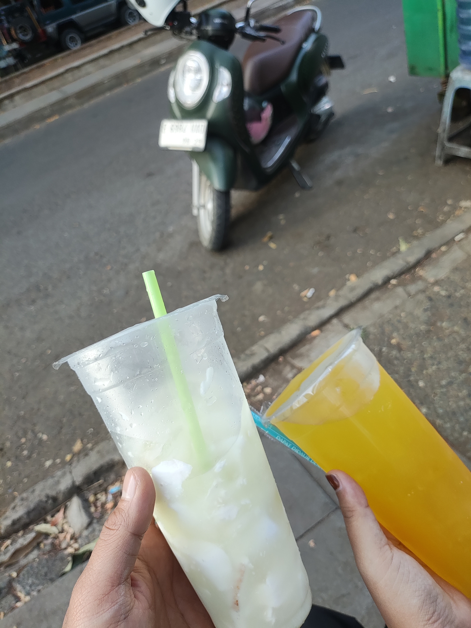 In Cirebon, Indonesia, two plastic cups of beverages are held by a person's hands, positioned in the foreground of the image. The cup on the left contains a light-colored, opaque liquid with visible white pieces, and a green straw. The cup on the right holds a bright yellow, translucent liquid. In the background, a dark green scooter is parked on a paved street. A dark-colored SUV is visible further in the background to the left. The environment appears to be an outdoor setting, possibly a roadside, with a sidewalk and some debris on the ground.