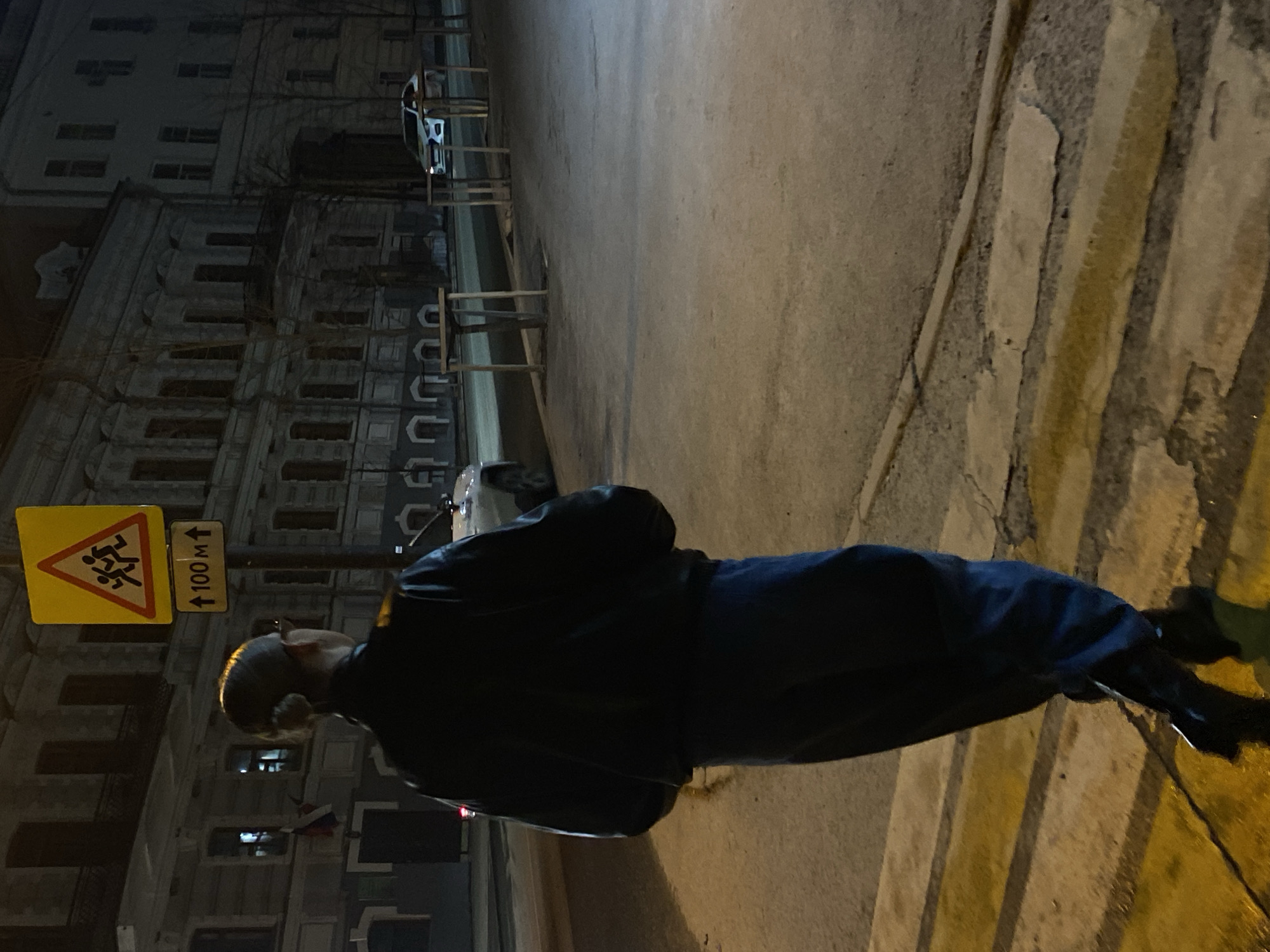 A person in a black jacket and dark pants is walking across a zebra crossing at night. Beside the person is a yellow triangular road sign depicting children running, with a sign indicating "100m." A light-colored, multi-story building lines the background, and a parked car is visible along the street. The location is likely Kazan, Russia, based on context clues.
