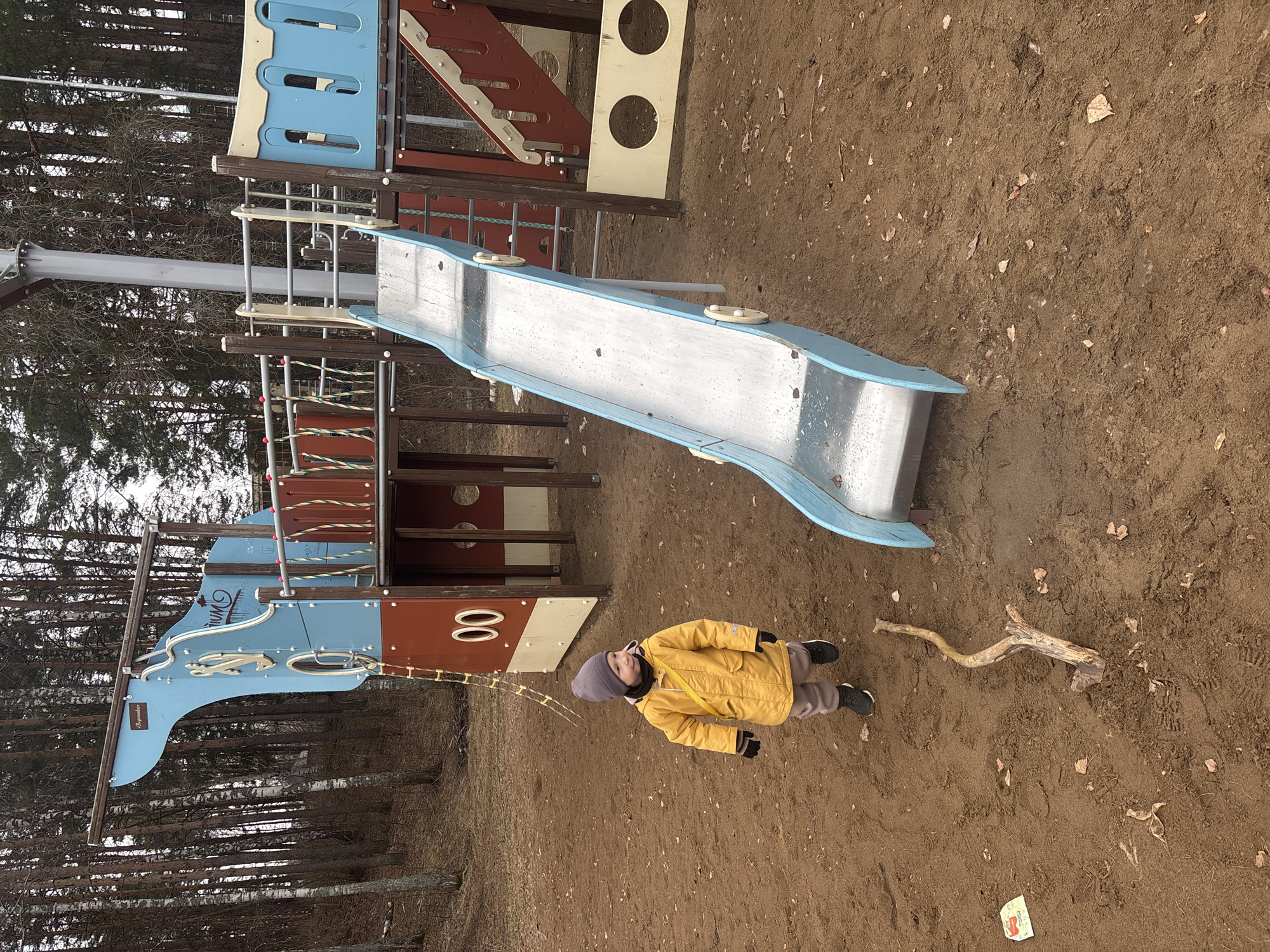 A child in a yellow jacket and grey hat stands on a brown dirt surface near playground equipment consisting of a metal slide and multi-tiered wooden structure, located in the city of Chaikovsky, Russia. A large branch lies on the ground near the child, and a small piece of trash is visible in the lower right corner of the image. Trees are visible in the background.