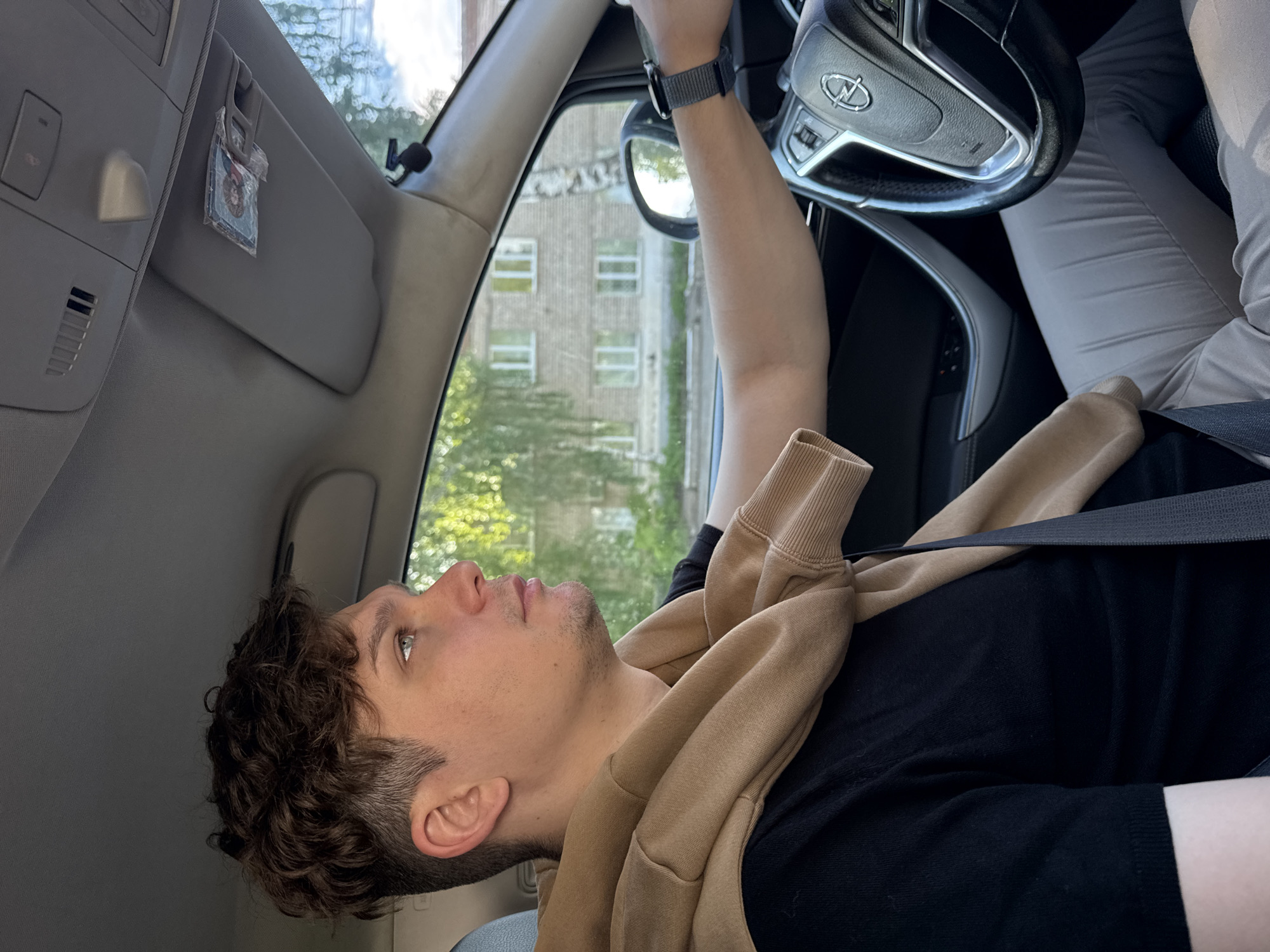 A young man is seated in the driver's seat of a car, looking out the window upwards and to his left. He has short, curly brown hair and a light stubble, with a neutral and contemplative expression. He is wearing a black t-shirt, with a light brown or beige sweatshirt draped over his shoulders. A black seatbelt crosses his chest. His right arm is visible, resting on the steering wheel, and he wears a dark grey smartwatch on his wrist.
The car's interior is light grey, with a sun visor on the ceiling from which a small, wrapped item (possibly an air freshener or ornament) hangs. The steering wheel, which bears a distinct Opel logo, is partially visible. The legs of another person, clad in light grey trousers, are seen in the passenger footwell.
Outside the car, through the window, an urban or suburban scene is visible. A multi-story brick building with multiple windows can be seen, partially obscured by green trees, suggesting a daytime setting with clear weather. The background indicates a setting in Chaikovsky, Russia. The overall impression is a moment of pause or observation while inside a vehicle.