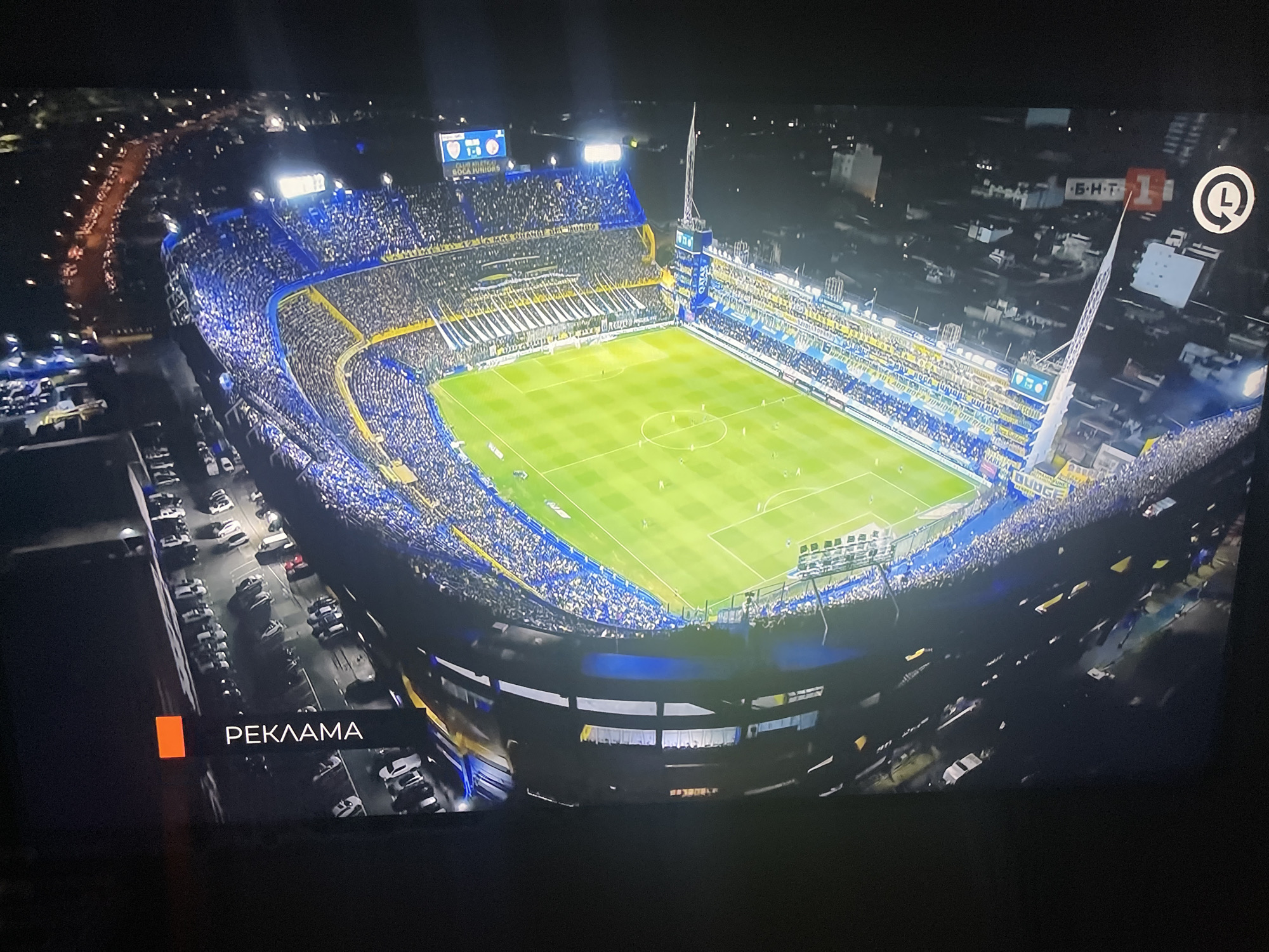 The image is an aerial view of a brightly lit football stadium at night, filled with spectators. The stadium features green turf with white markings and is surrounded by tiered seating filled with a crowd wearing predominantly blue and yellow. There are advertising banners visible around the stadium and a large scoreboard displaying "BOCA JUNIORS" and a score of "1-0". The stadium is situated within a city environment, with lights from surrounding buildings and roads visible in the background. A road with parked cars is visible in the foreground to the left, and a black bar at the bottom of the screen displays the text "РЕКЛАМА" (which means "ADVERTISEMENT" in Russian) alongside an orange rectangular icon. The scene depicts a sports event in progress. While the prompt mentions Ruse, Bulgaria, there is no information within the image itself that directly supports or denies this location.