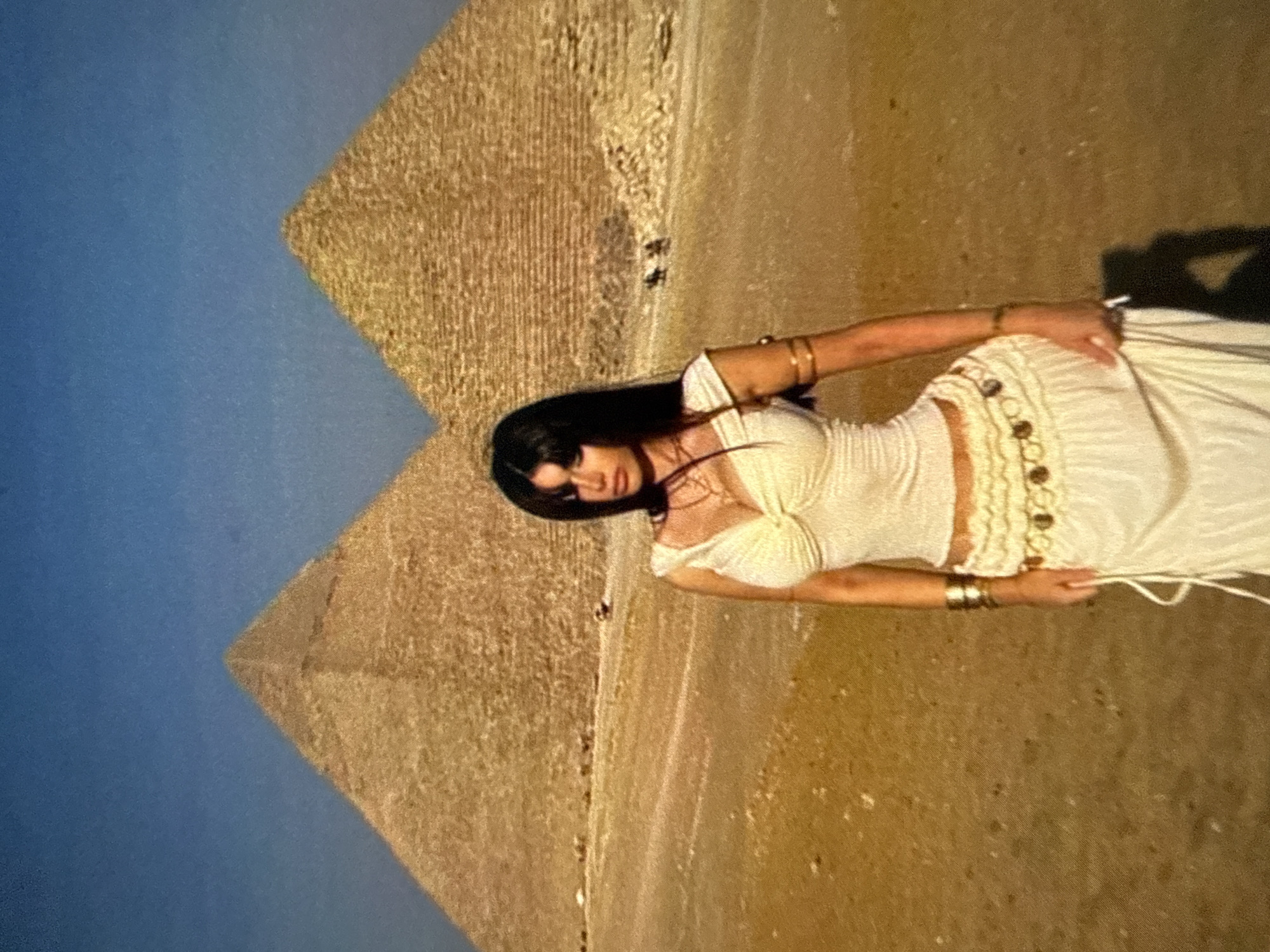 A woman in white clothing is standing in front of pyramids against a blue sky. The location is Egypt near the pyramids.