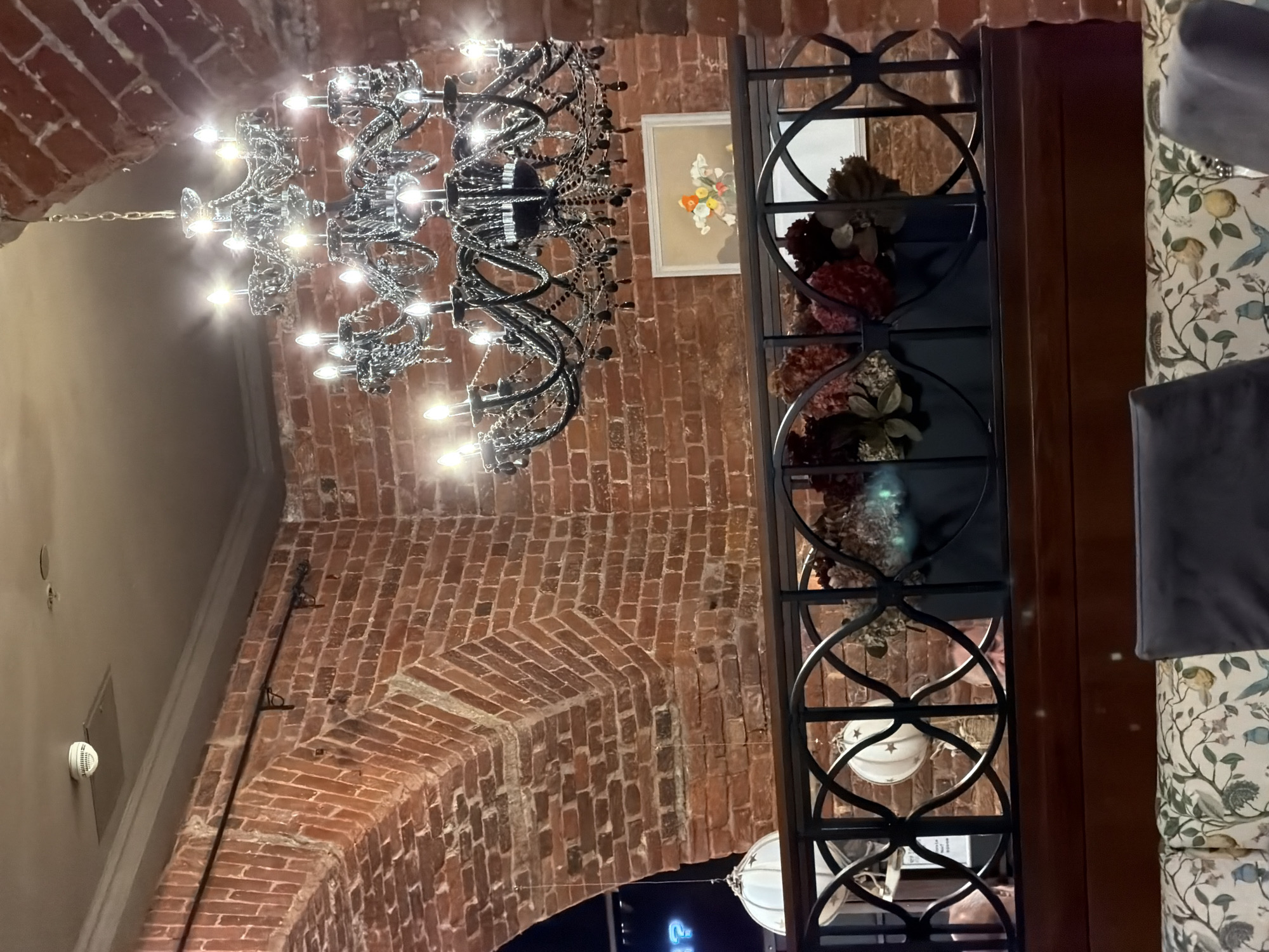 The image shows an interior scene, likely in a restaurant or cafe, with exposed brick walls and a large black chandelier hanging from the ceiling. A decorative metal railing with circular patterns is visible in the mid-ground, behind which are floral arrangements in dark rectangular planters. Above the railing, a framed artwork depicting flowers is mounted on the brick wall. In the background, a rounded brick archway is present. On the ceiling, a smoke detector is mounted. The foreground shows the tops of upholstered seating with a patterned fabric. The overall impression is of a rustic yet elegant setting. This image was taken in Saint Petersburg, Russia.