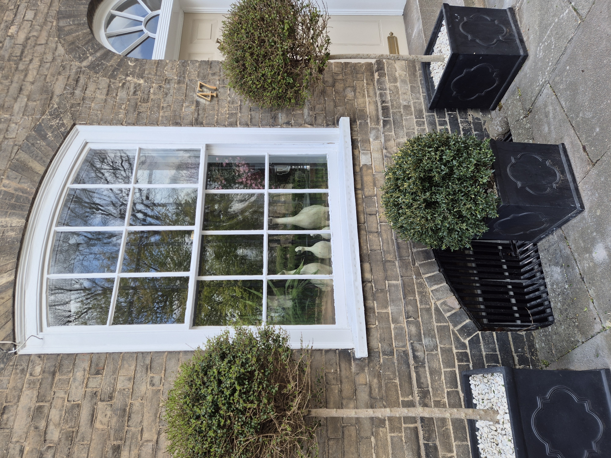 The image shows a section of a building exterior, likely a residential property, in West Suffolk, United Kingdom. The building is constructed of grey brick, with a large, arched window dominating the view. The window frame is white and reflects trees and the sky. Within the window are decorative objects resembling white swans. Above the window, near the front door, is the number '47'. Flanking the building are topiary bushes in black square planters, some containing white stones on top of the soil. The background and precise location within West Suffolk are not discernible from the image.