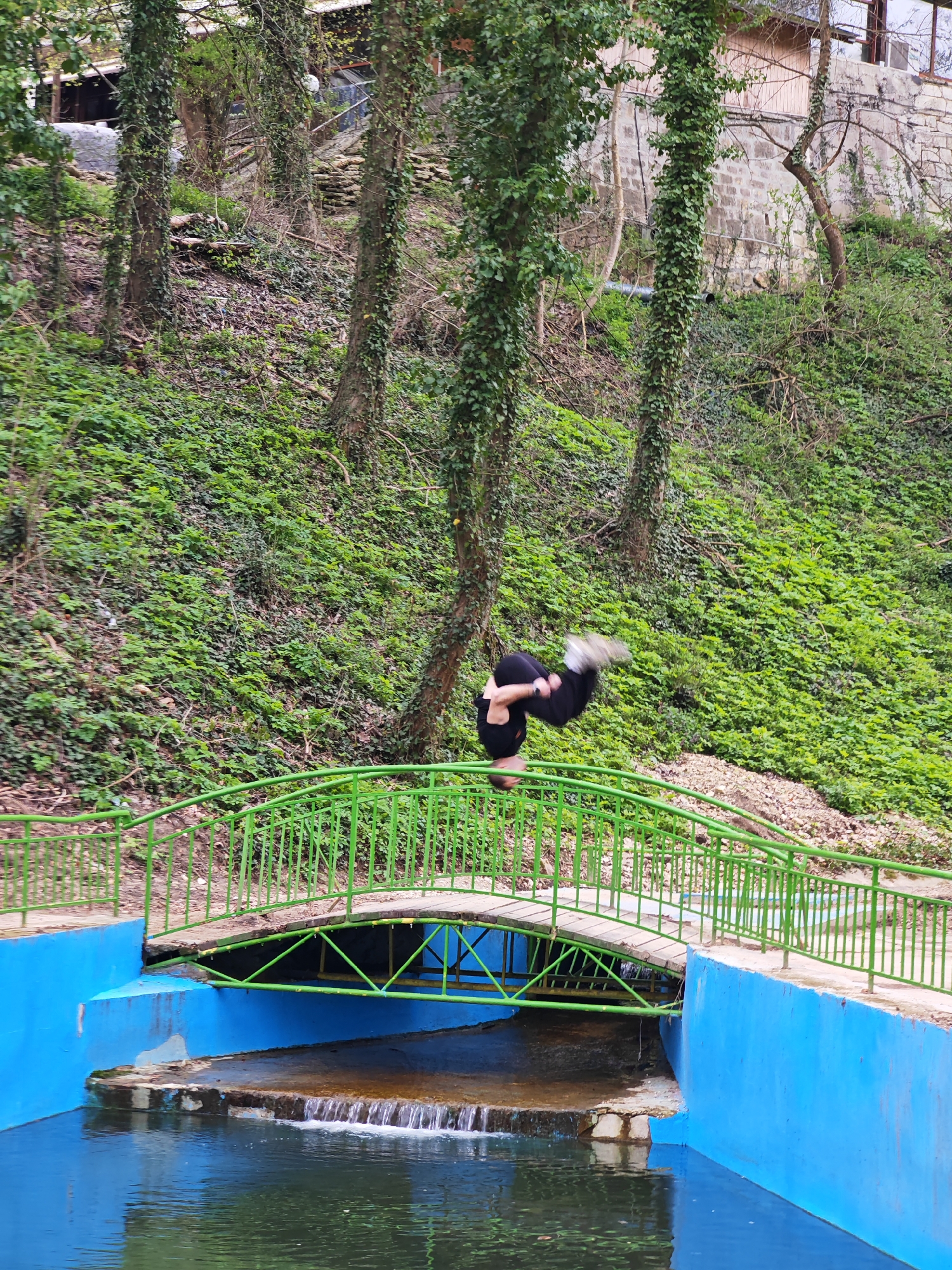 A person is in the middle of a backflip on a green bridge over a small canal with blue walls. The person is wearing dark clothing and light-colored shoes. The canal flows into a body of water. The background consists of a steep hillside covered in green vegetation and trees. This scene appears to be located in the city of Shumen, Bulgaria.