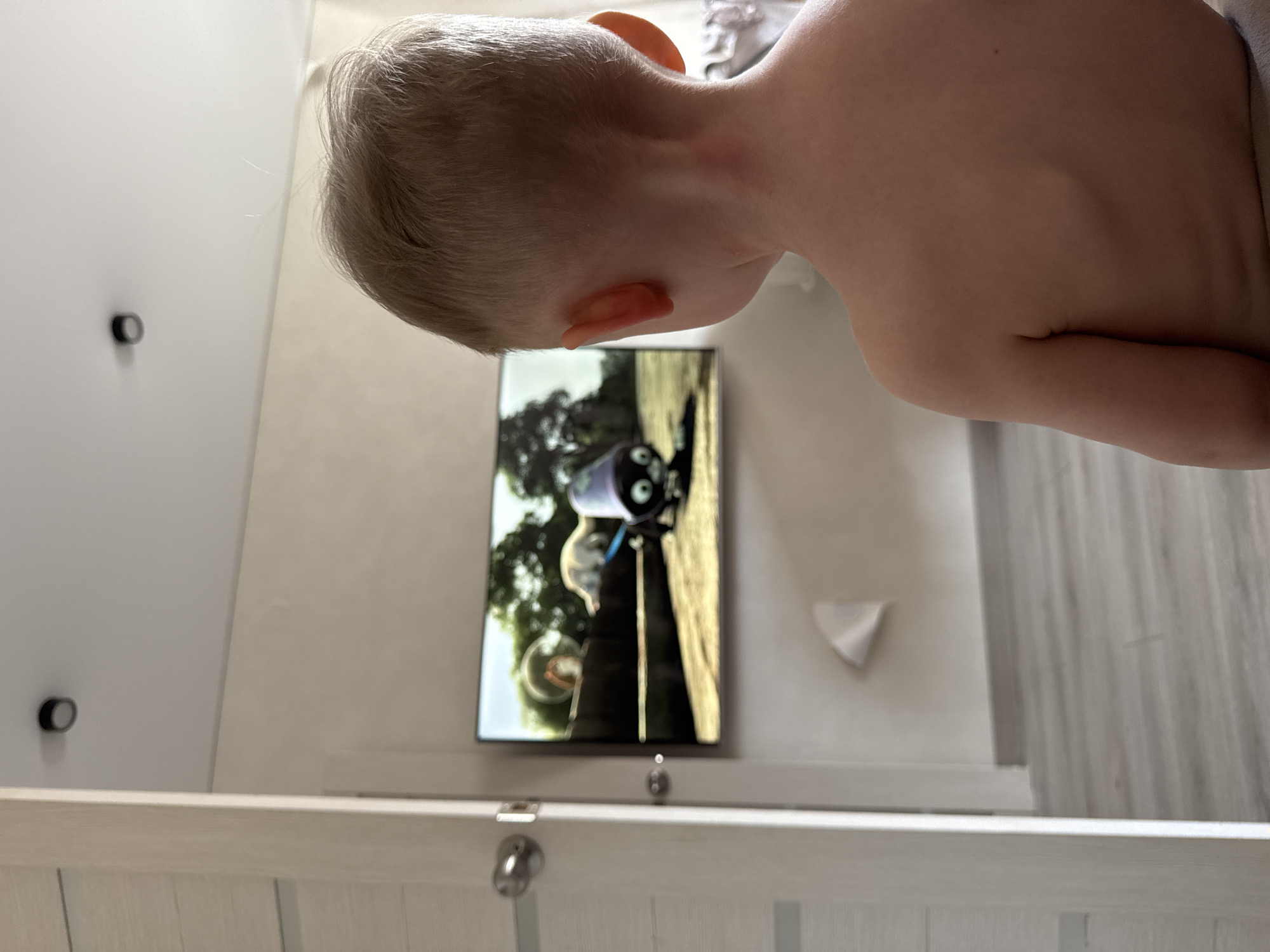 A young child, viewed from the back and side, watches a television screen. The child has fair hair and is shirtless. The television displays an animated scene featuring what appears to be a robot or character with large eyes, set against a background of trees and a golden path. The setting is indoors, likely a bedroom, with a crib or bed in the foreground. The walls are a light color, and there are two black circular fixtures on the left wall. A white triangular object is affixed to the wall on the right. The lighting suggests it is daytime. No text is visible. The overall mood seems relaxed and focused, with the child engrossed in the program.