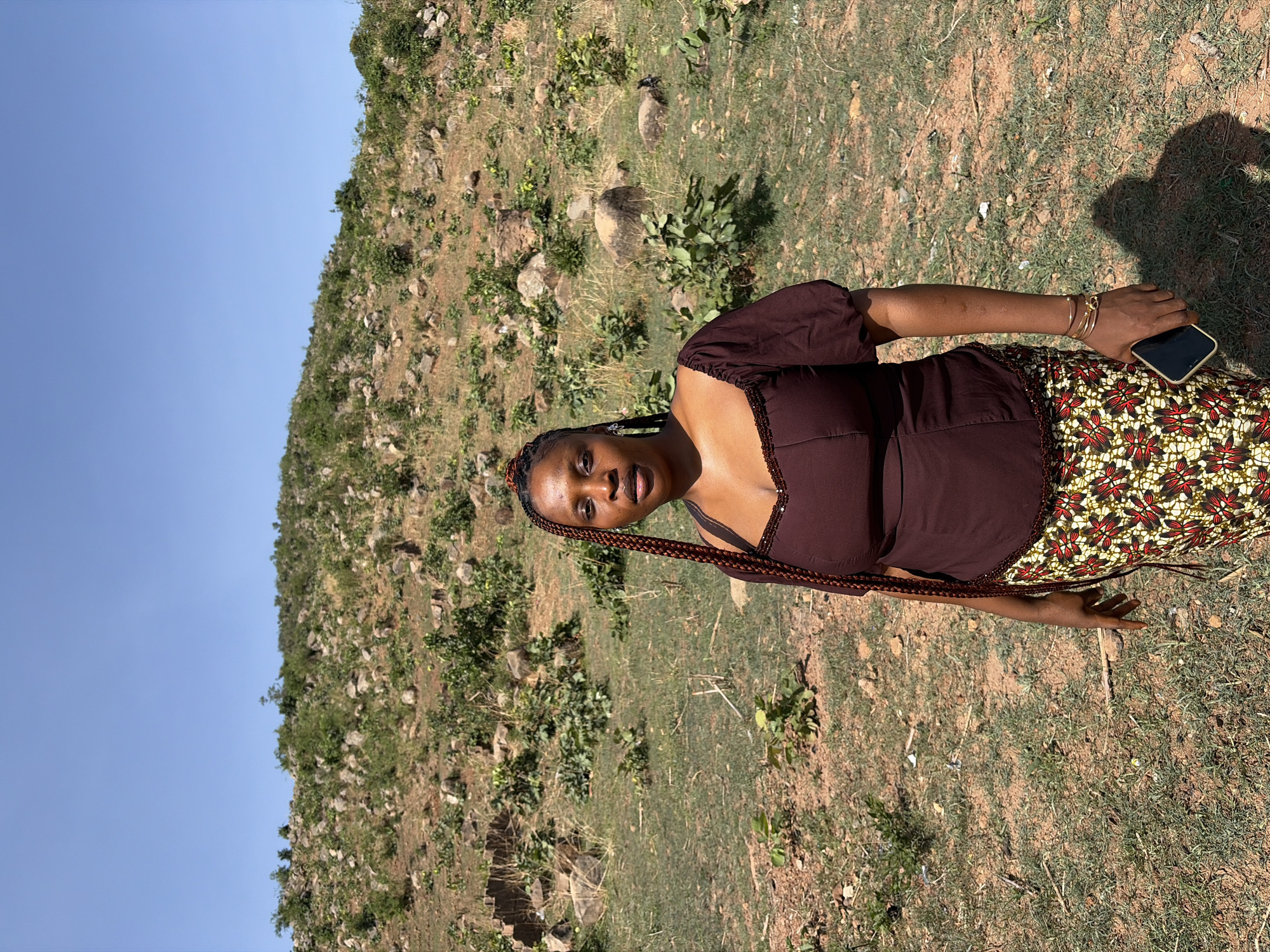A young woman with dark skin and braided hair stands in an outdoor setting. She is wearing a brown, short-sleeved blouse and a skirt with a colorful, repeating floral pattern. She holds a cell phone in her right hand. The background consists of a hill or mountain covered with sparse vegetation, rocks, and brown earth, with a clear blue sky above. The scene suggests a natural environment, potentially a rural or semi-rural area outside an urban setting. The location is the city of Jos, Nigeria.