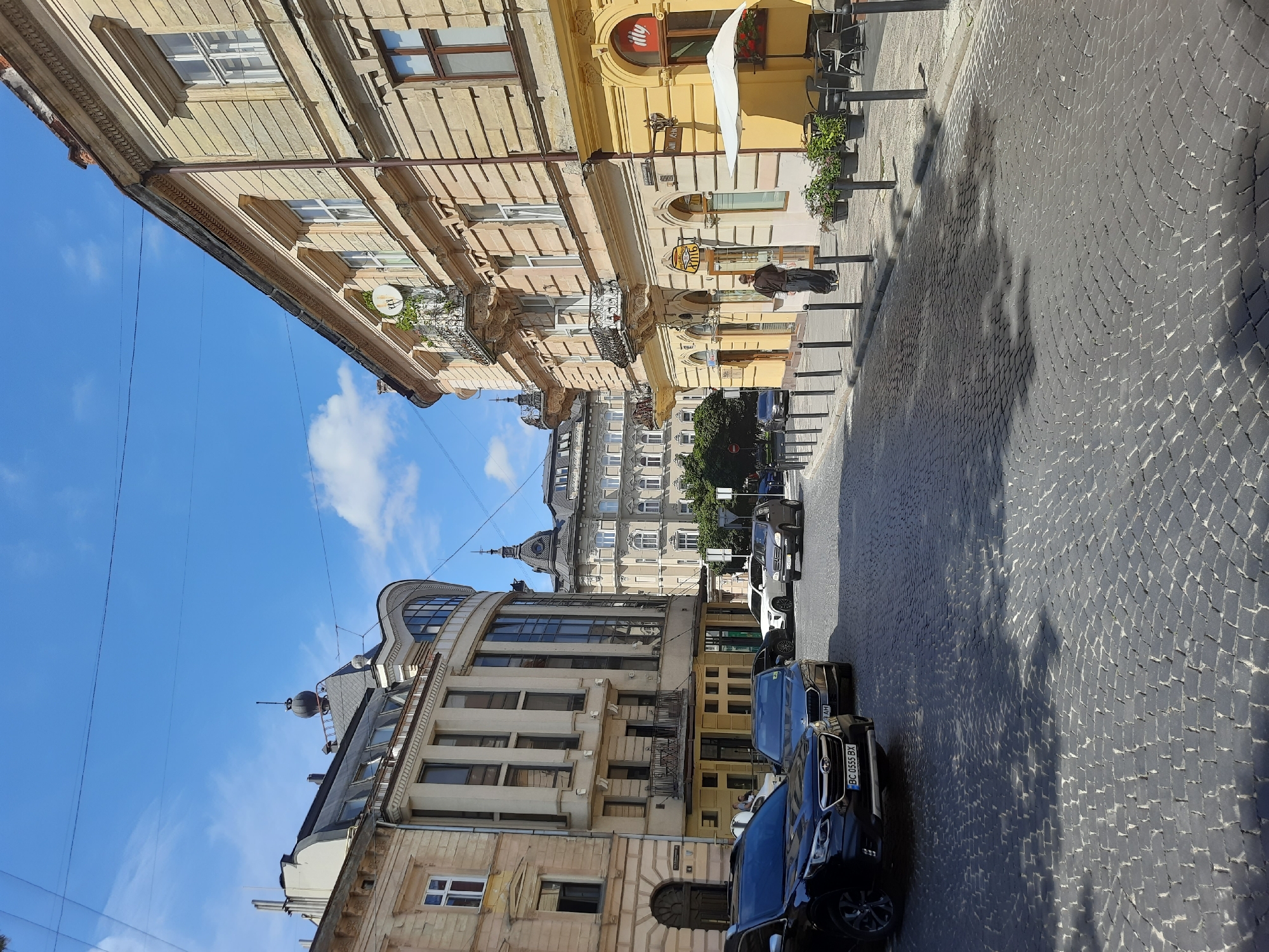 The image depicts a sunlit street scene in Lviv, Ukraine. Cobblestone paving covers the street and sidewalk. On the right side of the street, a pale yellow building features a cafe with outdoor seating under a white umbrella, marked by the "illy" logo. The cafe's windows display signage. A single person, dressed in dark clothing, walks along the sidewalk. Several cars are parked on the street, including a black SUV in the foreground with a Ukrainian license plate. Buildings with ornate architectural details line both sides of the street. The sky above is blue with scattered white clouds. The scene appears to be in a well-preserved historical district of Lviv.
