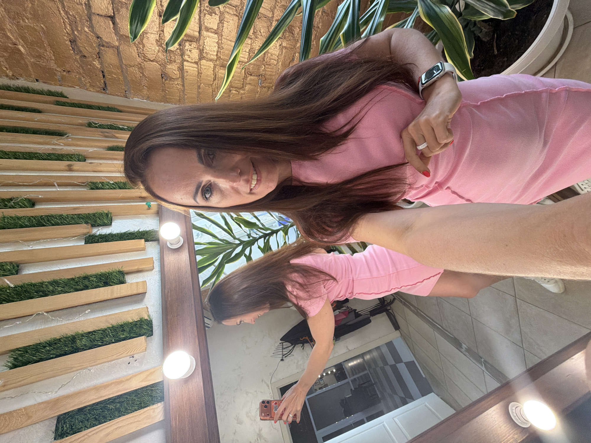 A woman is taking a selfie in front of a mirror. She is wearing a pink dress and a wristwatch, and she is holding a mobile phone. The background includes a wall with wooden planks and artificial grass, a brick wall, and a potted plant. The location is not discernible from the image and cannot be identified as Birobidzhan, Russia.