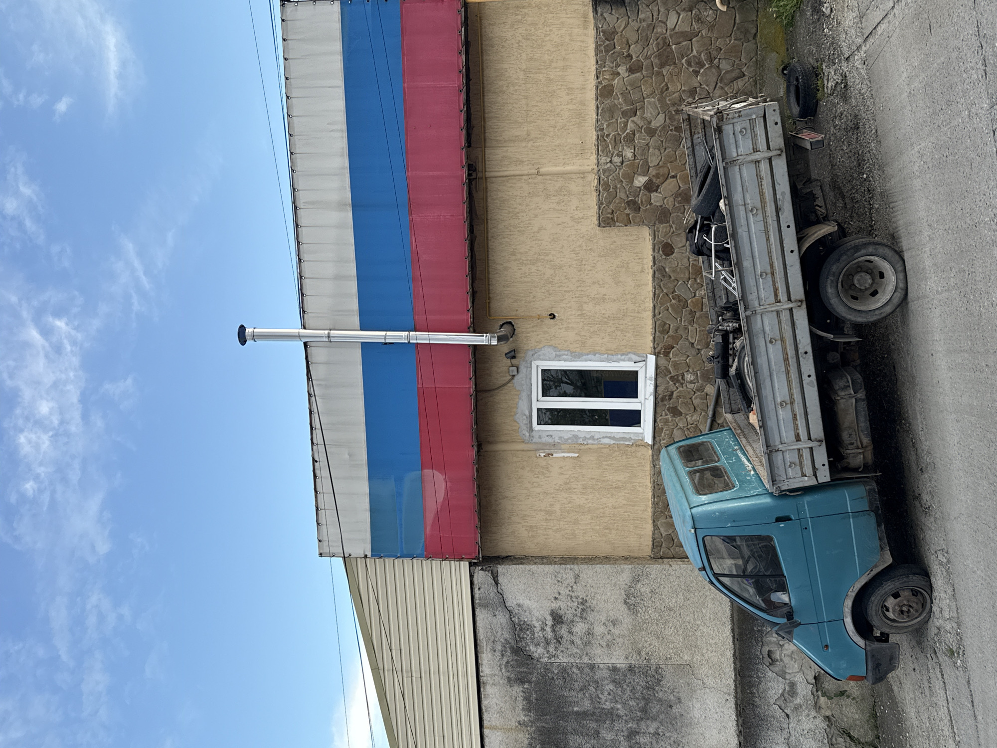 A partially occluded view of a building and a blue truck are shown. The building is adorned with a facade painted with the colors of the Russian flag. Adjacent to this section of the building is a wall covered in stone cladding and a window with a white frame. The truck appears to be parked alongside the building on what appears to be a road or driveway. The location is likely Sochi, Russia, given the color scheme of the building's facade.