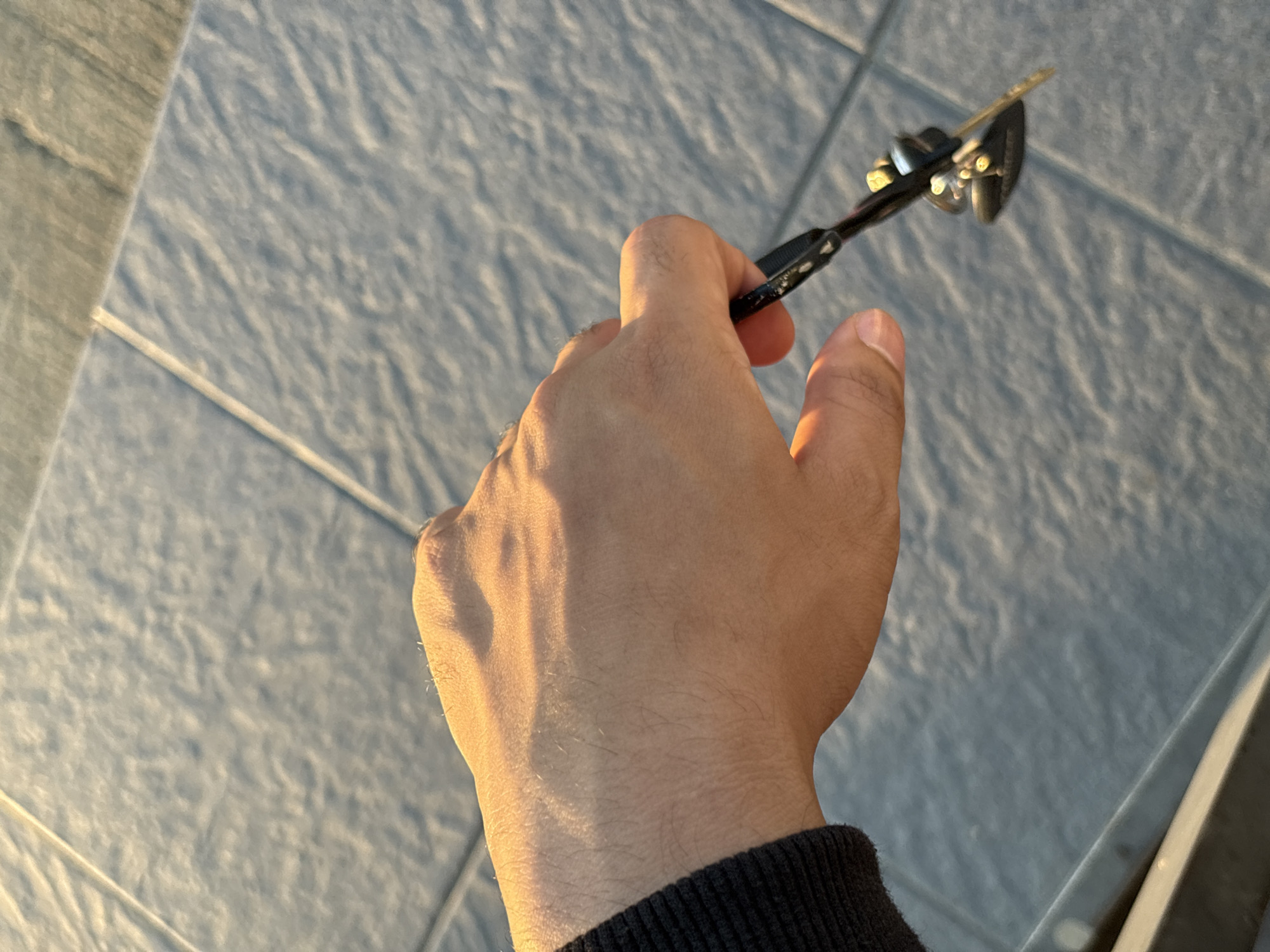 A hand, with visible arm hair and wearing a dark sweater, holds a pair of black eyeglasses with gold accents near the temple. The eyeglasses are angled downwards. The hand is positioned over a tiled surface that appears to be an outdoor pavement, with visible grout lines between the square tiles. Sunlight casts a warm glow on the hand and a portion of the tiled ground. The background is out of focus, showing more of the tiled surface. The setting appears to be a public space, possibly a plaza or walkway, and based on the context provided for Tagum City, Philippines, the environment is consistent with an urban outdoor area within the city.