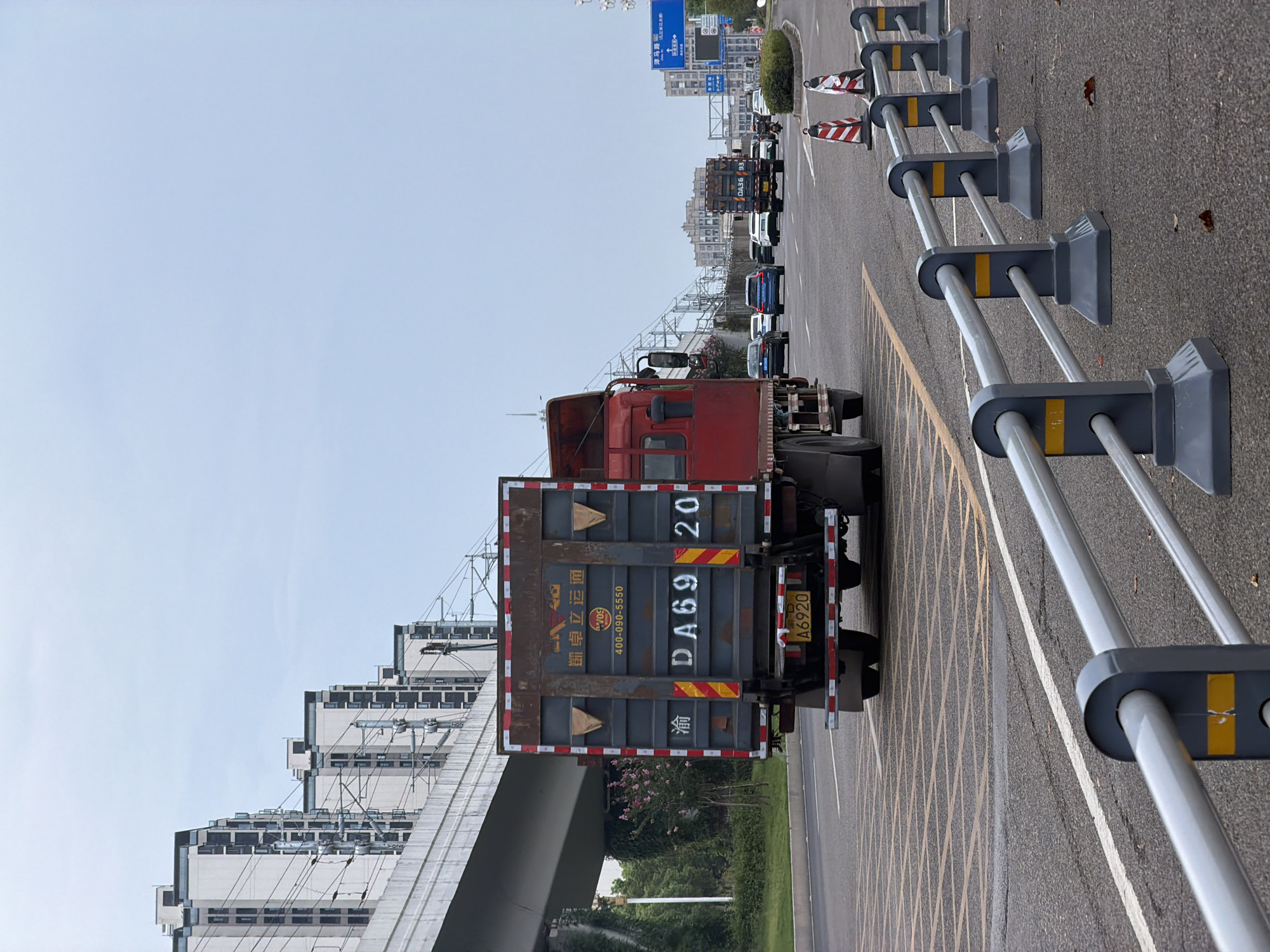 In Jiangjin, China, a red truck with a dark gray trailer is driving on a road. The trailer displays the license plate "DA6920" and Chinese characters. Other vehicles, including a blue car and a motorcycle, are also visible on the road. Buildings and a railway bridge are in the background. The road has yellow markings and a metal guardrail. The sky is overcast.