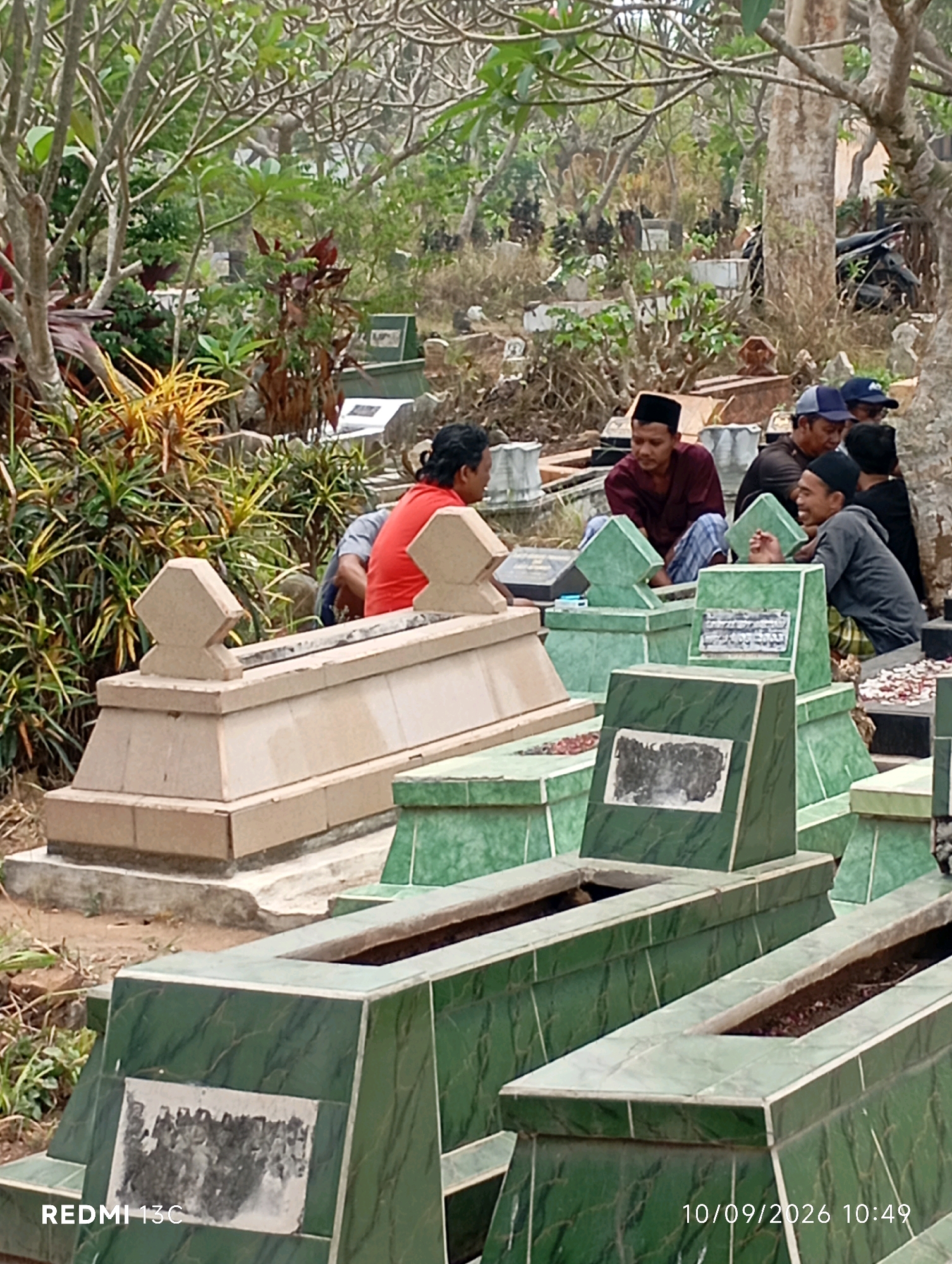 The image depicts a graveyard in Rambipuji, Indonesia, filled with numerous green and beige-colored tombstones. Several men, dressed in traditional Islamic attire including peci caps, are gathered around the graves, some sitting and conversing while others appear to be observing. The environment is characterized by lush green vegetation and trees, suggesting an outdoor, possibly tropical setting. The presence of multiple graves and the gathering of people indicate a communal or ceremonial occasion, likely a burial or a visit to pay respects. The overall atmosphere appears solemn yet communal.