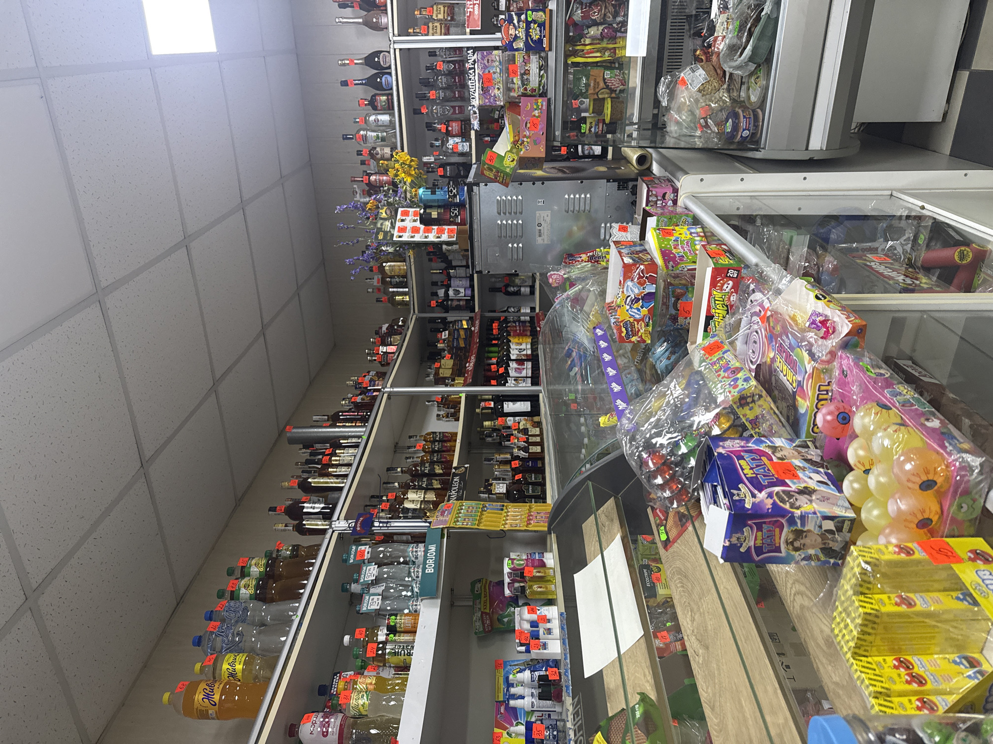 The image shows the interior of a small convenience store in Obukhivka, Ukraine. The store is filled with shelves stocked with various items, including beverages, snacks, and candies. On the top shelves, there are numerous bottles of water, juices, and alcoholic beverages, including several bottles of Borjomi mineral water. Various brands of liquor are displayed on the shelves, along with what appear to be wine bottles.
In the middle section of the store, there are displays of packaged food items and smaller bottles of liquids. There is also a large metal appliance, possibly a refrigerator or freezer unit, situated centrally.
The lower shelves and display cases are filled with a variety of candies, snacks, and smaller packaged goods. Some of these include colorful boxes of sweets, bags of chips, and other novelty items. A glass display case in the foreground contains more packaged snacks and what appear to be dried fruits or nuts. The store has a tiled floor and fluorescent lighting in the ceiling. The overall impression is of a well-stocked, typical small grocery or convenience store. There are no people visible in the image.