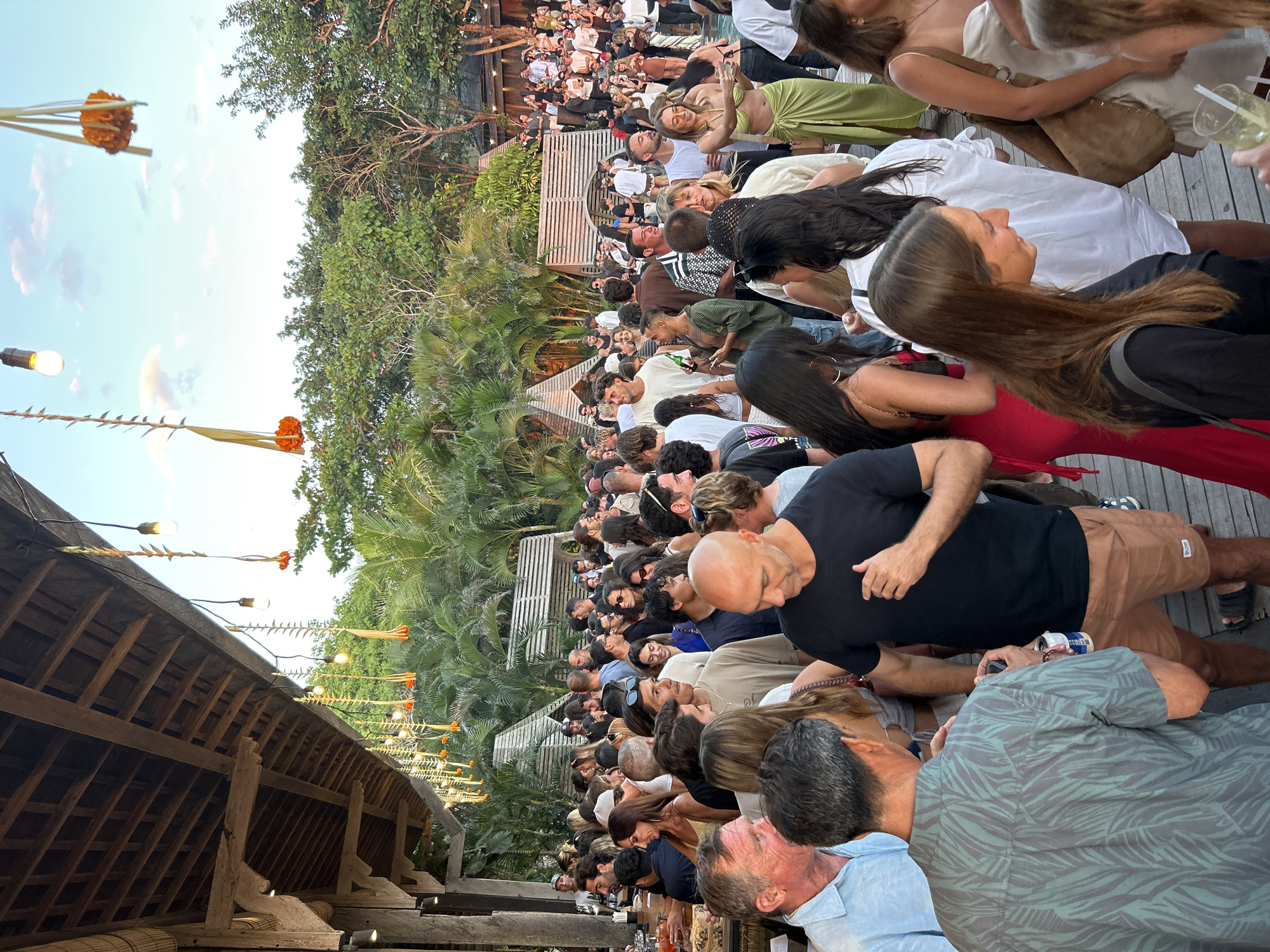 This image depicts a lively outdoor gathering, possibly a party or social event, at a location in Kuta Selatan, Indonesia. A large crowd of people, appearing to be young to middle-aged adults, are mingling. The setting is characterized by rustic wooden architecture, with an exposed beam ceiling and thatched roofing visible in the foreground. Decorative elements, including string lights and orange flowers interspersed with dried foliage, hang from the eaves. Lush green tropical trees and palm fronds surround the area, suggesting a warm climate.
Many individuals are dressed in casual summer attire, with some wearing swimwear or beachwear, like bikinis and shorts. People are engaged in conversations, holding drinks, and some appear to be dancing or moving through the crowd. The overall atmosphere seems festive and energetic. The ground appears to be a wooden deck or platform. In the background, there are glimpses of other structures, possibly seating areas or pavilions, also built with natural materials. The sky is visible above, with some clouds present, indicating it is either late afternoon or early evening.