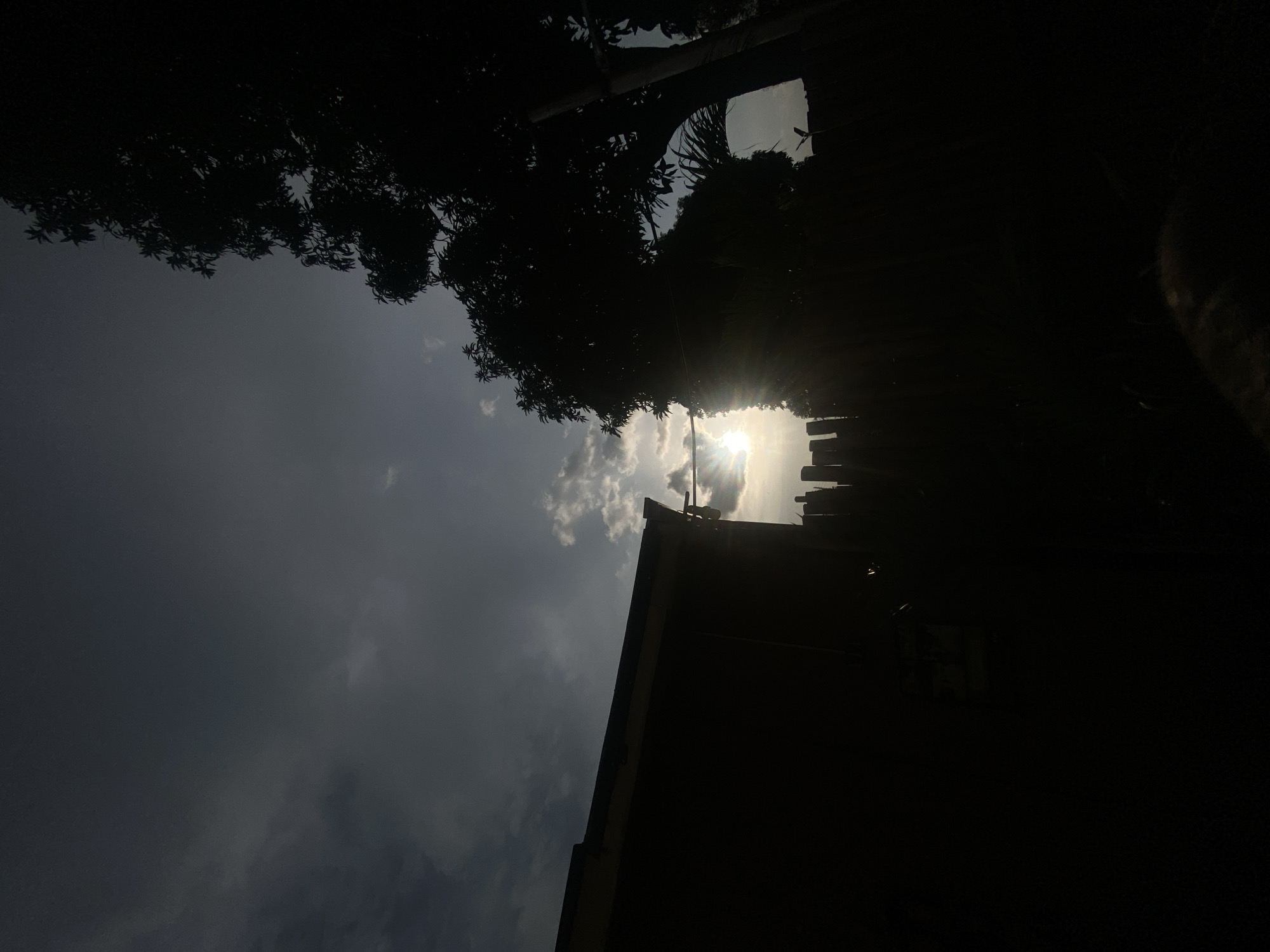The image captures a silhouetted scene of the sky and the outlines of natural and man-made structures, with the sun breaking through clouds and casting lens flares. The location is identified as Barden Yana, Nigeria.
In the foreground, on the left, is the dark silhouette of a building, possibly a house, with a visible roofline. To the right of the building, a fence made of vertical wooden posts is partially visible, also in silhouette. Behind the fence, a large tree with dense foliage dominates the right side of the frame, its branches and leaves creating a dark, irregular shape against the sky. A thin wire or cable stretches horizontally across the mid-ground, passing in front of the tree.
The sky is a prominent feature, with a gradient of dark grays and blues. Patches of lighter clouds are interspersed, and the sun is positioned just above the fence line, its bright light causing significant lens flares that spread across the image, particularly around the sun itself and towards the left side. The overall lighting suggests either sunrise or sunset.
There are no people or visible actions taking place. The environment appears to be an outdoor setting, likely a residential area given the presence of the building and fence. The silhouette effect and the dramatic lighting create a moody and atmospheric composition. The scene is static and captures a specific moment in time.