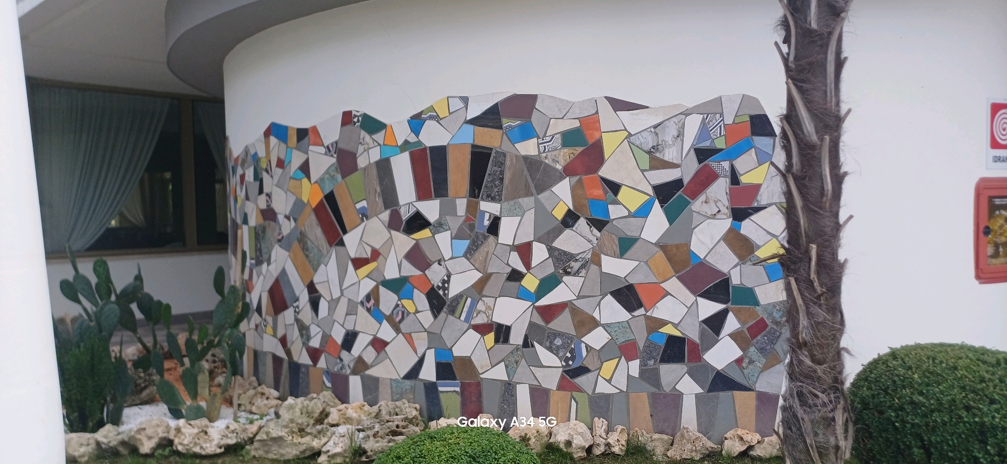 The image shows an exterior view of a building with a curved white wall adorned with a colorful mosaic. The mosaic is made of irregularly shaped pieces of tile in various colors and patterns, including geometric designs, solid colors like red, blue, yellow, and green, and marbled textures. To the left of the mosaic wall, there are several prickly pear cacti and some greenery. A tall, thin palm tree trunk with a rough, fibrous texture stands to the right of the mosaic. Behind the mosaic, a white wall continues, with a window covered by light blue curtains visible. A small red framed object with a circular target-like symbol and the word "IDRAN" is mounted on the white wall to the right of the palm tree. In the foreground, there are rocks and lush green ground cover. The overall scene suggests an outdoor area of a resort or a public building in a warm climate. The "Galaxy A34 5G" watermark in the lower right corner indicates the device used to capture the image. This image is likely taken in Abano Terme, Italy, known for its thermal spas and hotels.