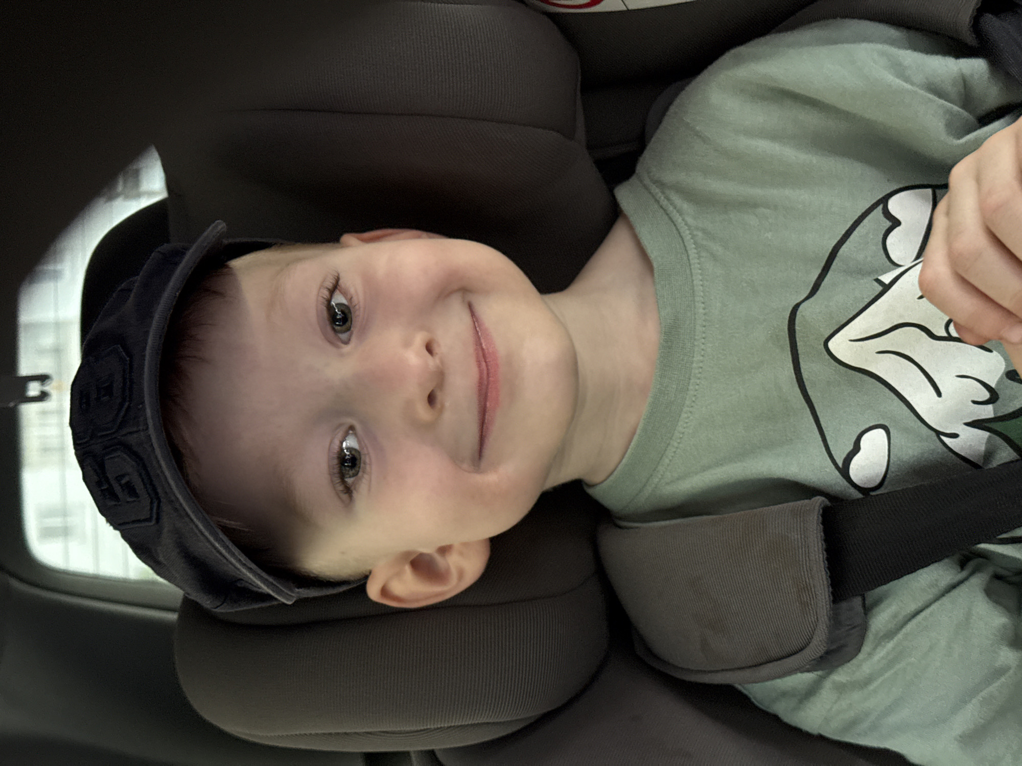 A young child, presumably male, is securely fastened in a car seat. The child is wearing a dark cap and a light green shirt with a white and black design. The child is looking directly at the camera and smiling slightly. The car seat is dark gray. The location is not discernable based on the image.