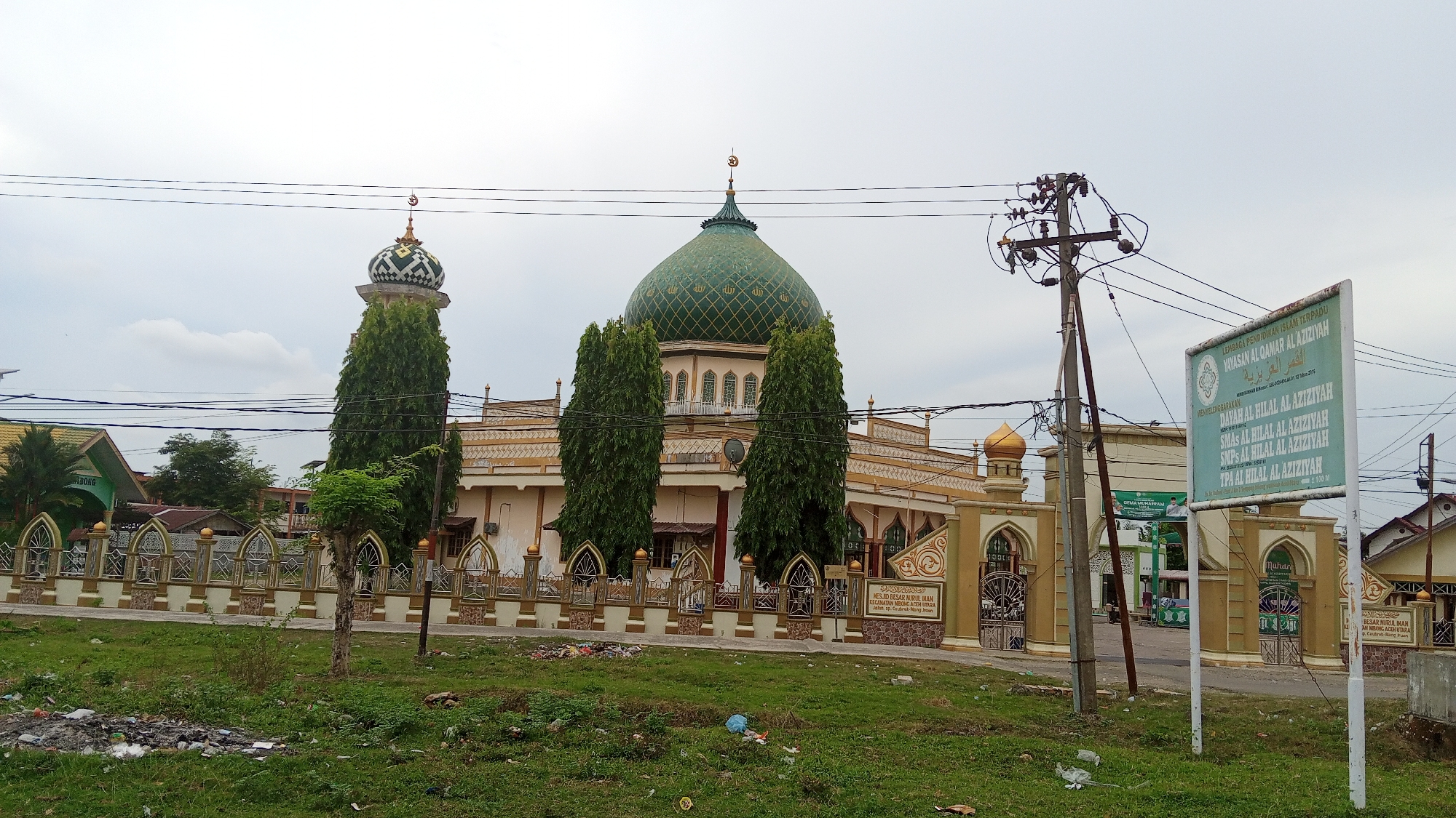 The image shows a mosque in Keudenibong, Indonesia. The mosque has a prominent green dome and a tall minaret. A fence with decorative elements surrounds the mosque complex. A sign on the right side of the image displays text in Arabic and Indonesian, indicating it is associated with an Islamic educational foundation. In the foreground, there is a grassy area with some scattered litter. Power lines and a utility pole are visible in the background. The sky is overcast.