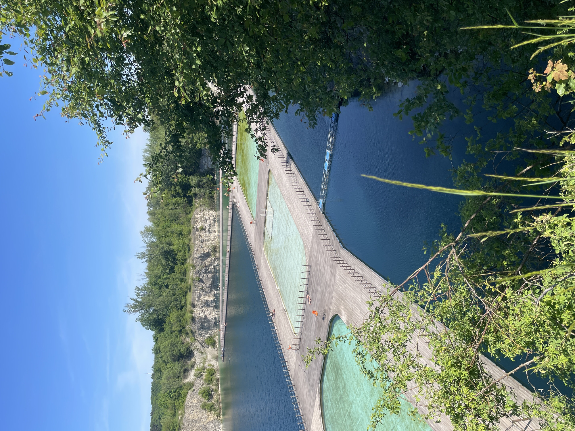 The image shows an aerial view of the Blågårdsgade Bad floating swimming pool in Copenhagen, Denmark. The pool is located in a former limestone quarry, with a series of wooden walkways connecting different sections of the pool. The water in the pool appears to have varying shades of blue and green, likely due to depth and algae. Some people are visible sunbathing on the wooden walkways. The surroundings include a rocky cliff face covered with trees and vegetation. The sky is blue with some clouds.