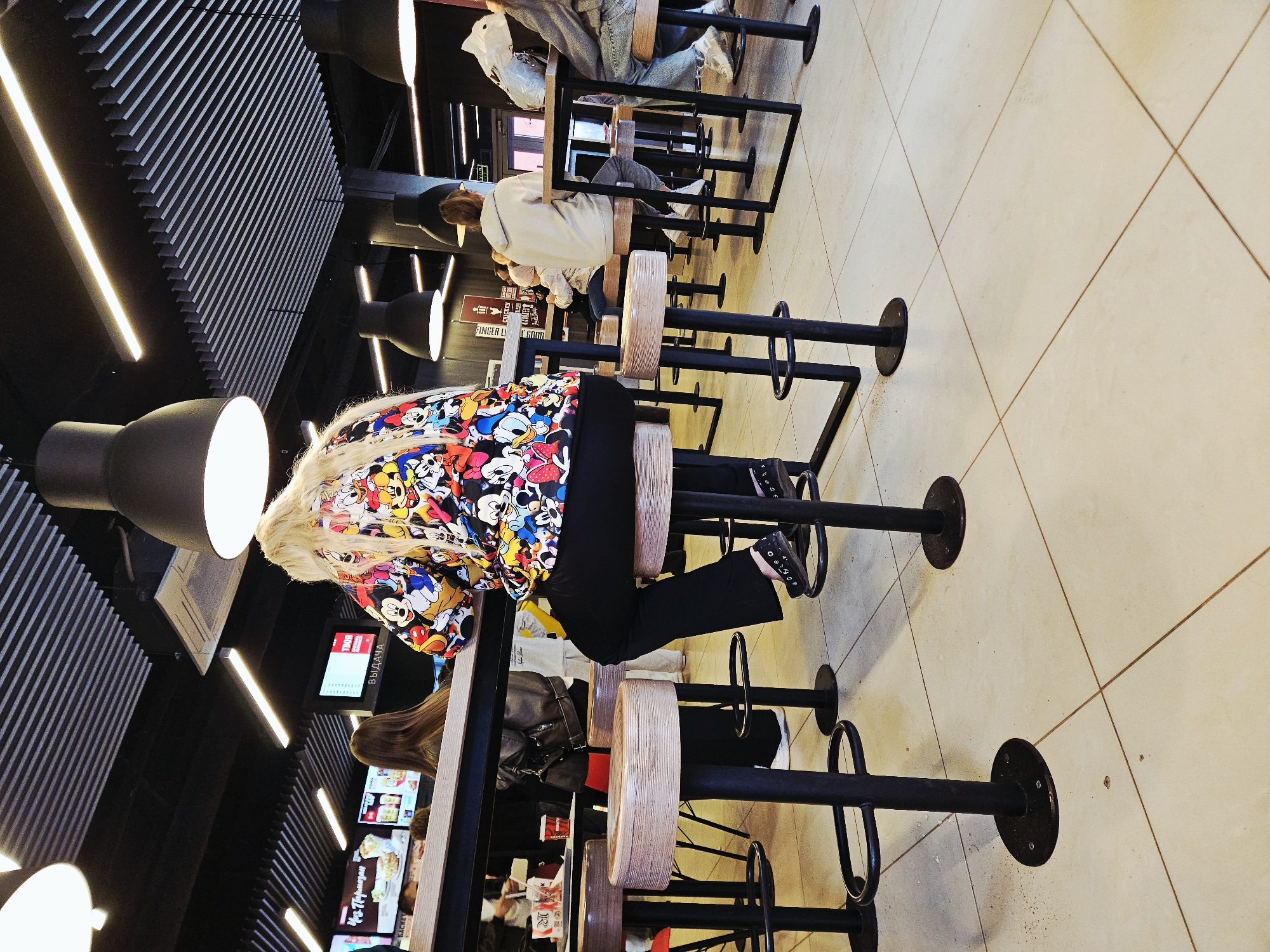 The image depicts the interior of a fast-food restaurant in Ulyanovsk, Russia, likely a KFC based on signage visible in the background. The scene is set at a high-top table with stools. In the foreground, a woman with long blonde hair sits facing away from the camera, wearing a colorful jacket featuring cartoon characters, black pants, and black shoes. She is seated on a stool at a wooden table. Several other individuals are visible in the background, some seated at tables and others appearing to be in line. The restaurant's interior features black ceilings with modern lighting fixtures and a tiled floor. Menu boards are visible behind the counter area.