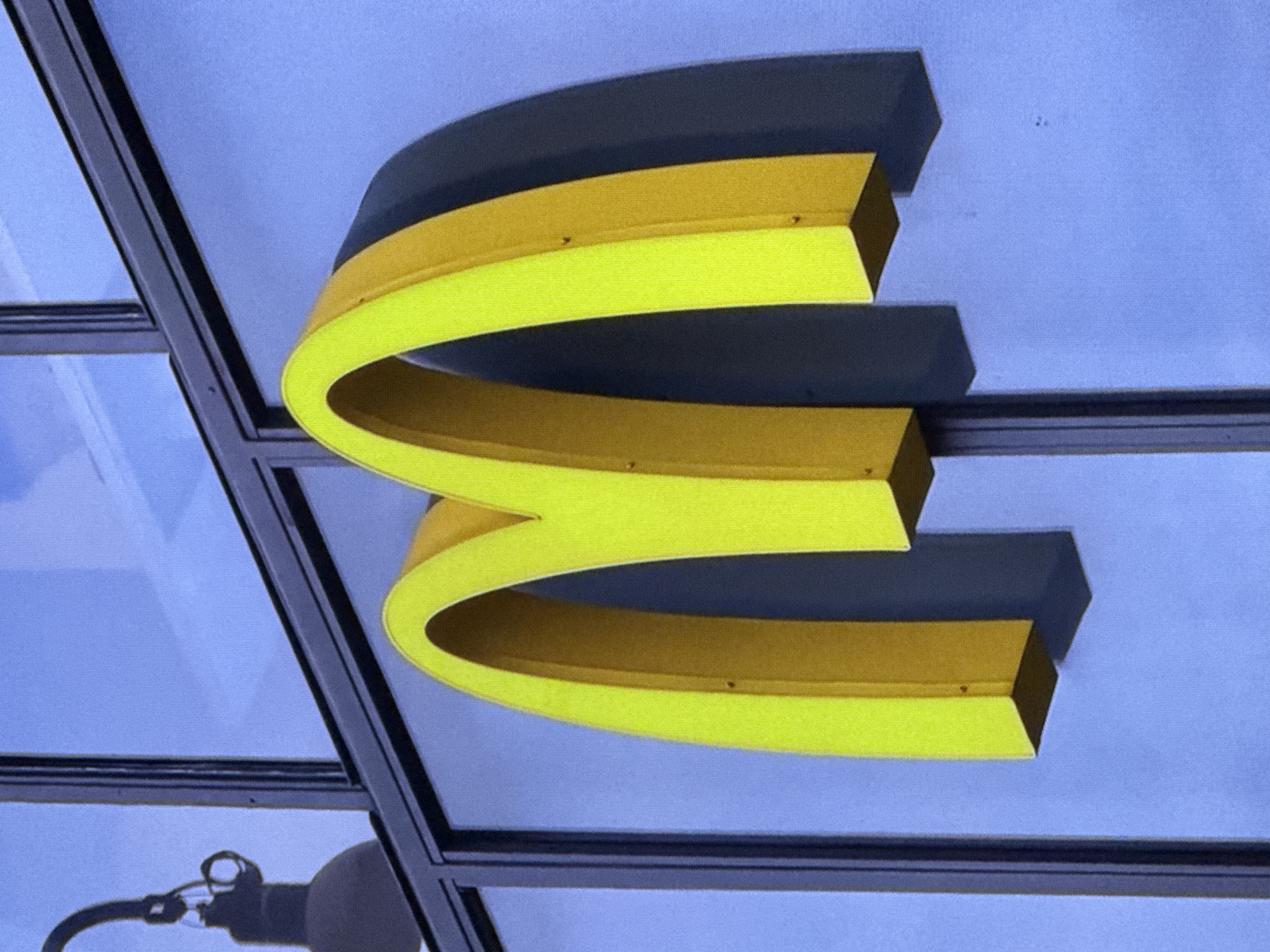 The image shows a close-up of the iconic golden arches of a McDonald's restaurant sign, rendered in bright yellow. The sign is mounted on a building with a large glass facade, reflecting a clear blue sky. A dark shadow of the arches is cast on the glass behind them, indicating the direction of the light source. A light fixture, partially visible in the upper left corner, is also reflected in the glass. The sign appears to be a freestanding or exterior mounted sign, typical of fast-food establishments. The overall impression is one of clear daylight and modern architecture.