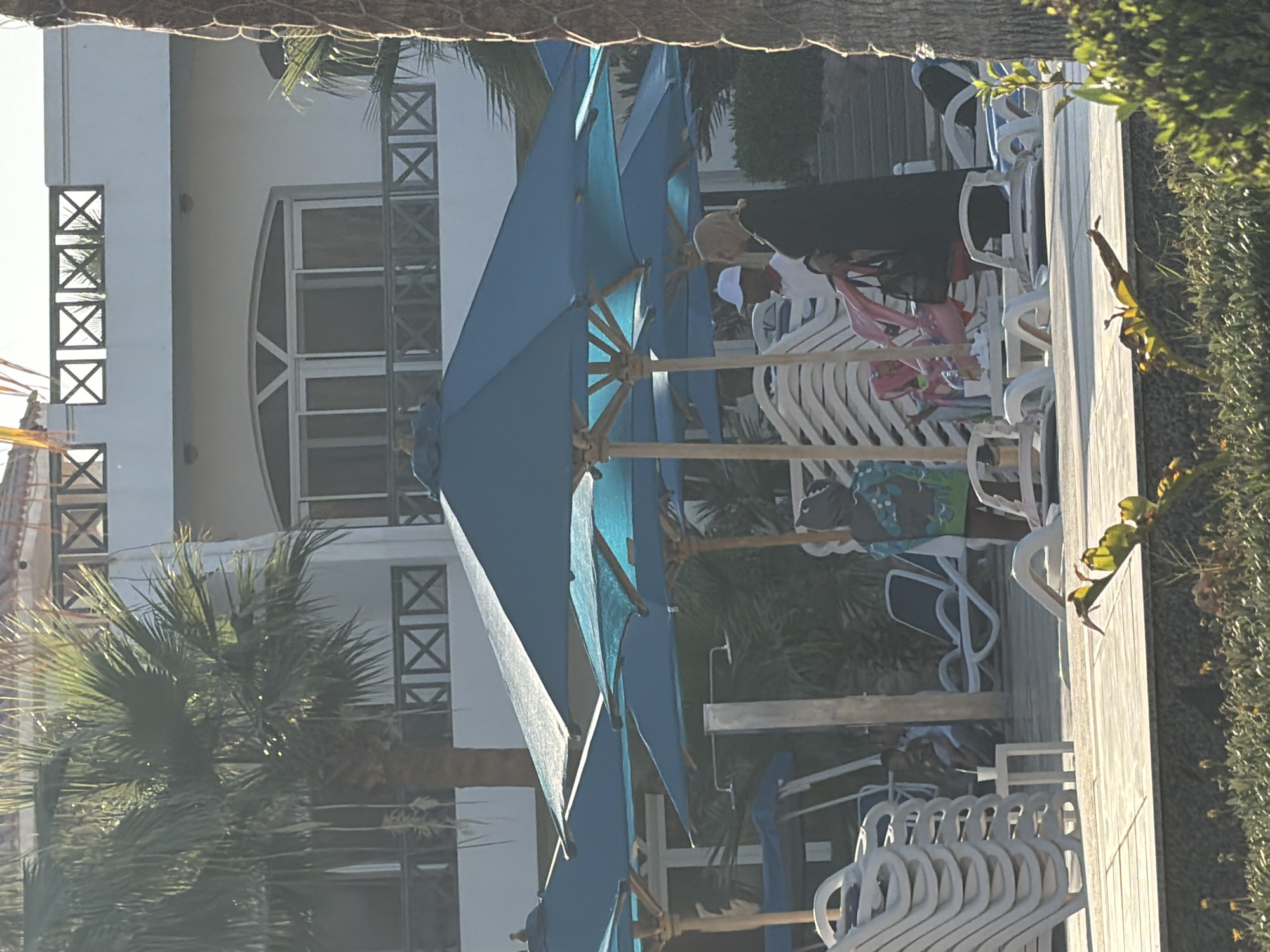 In Sharm El Sheikh, Egypt, an outdoor scene unfolds near a resort. Several large, teal-colored umbrellas provide shade over a wooden deck. Stacked white lounge chairs are visible, suggesting a poolside or beachfront area. In the mid-ground, three individuals are standing under one of the umbrellas. One person, wearing a white baseball cap and a white shirt, is facing away from the camera. Next to them, a person wearing a light-colored hijab and a dark abaya is also standing. Between them, a person in a dark hooded robe with a green and blue pattern is partially obscured. In the background, a multi-story white building with balconies and latticed railings is visible, surrounded by palm trees and lush greenery. The overall impression is a relaxed atmosphere in a tropical resort setting.