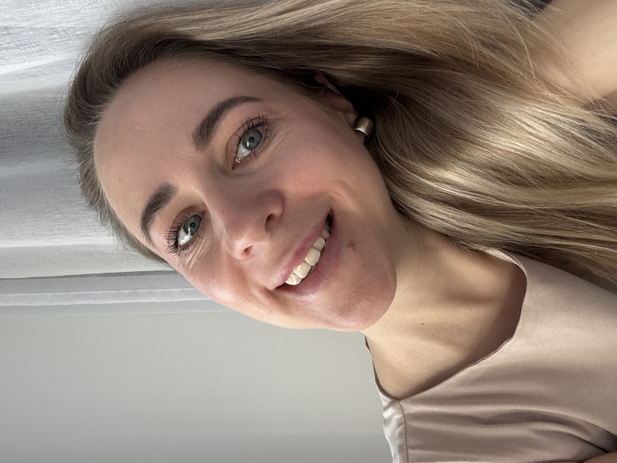 A Caucasian woman with blonde hair and blue eyes smiles at the camera. She wears a light pink or beige colored top. She has a silver hoop earring in her left ear. The background appears to be a white ceiling or wall. There is no evidence to suggest this image was taken in Chaikovsky, Russia.